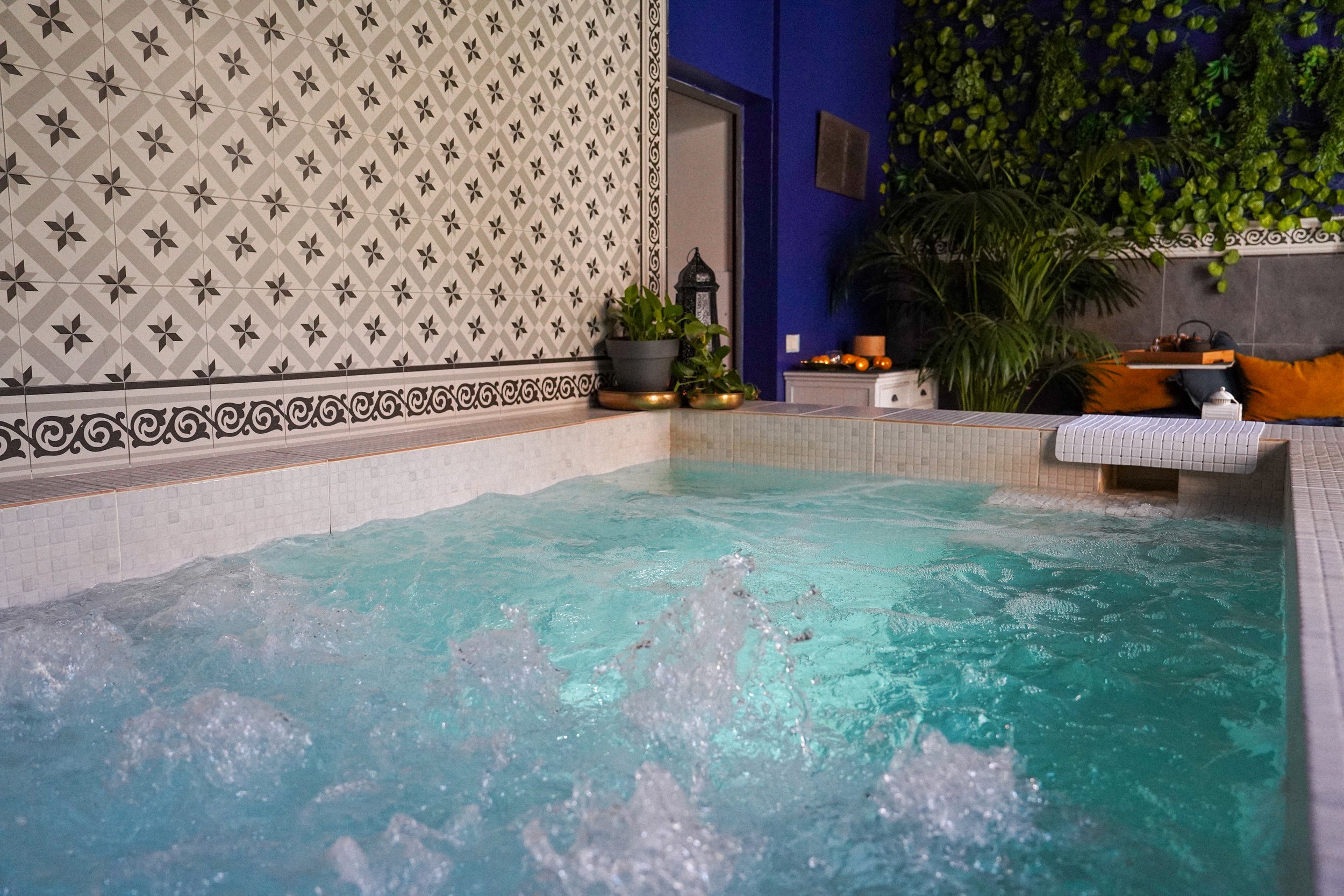 Piscina con agua caliente y azulejos decorativos en la pared, rodeada de plantas verdes y cojines naranjas.