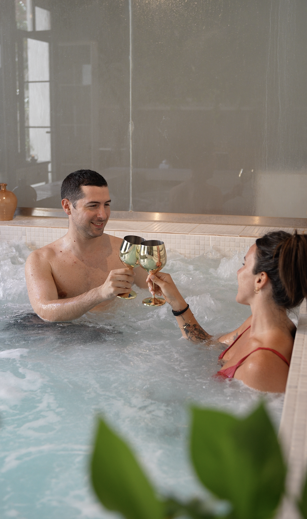 Una pareja en una bañera de hidromasaje brindando con copas doradas.