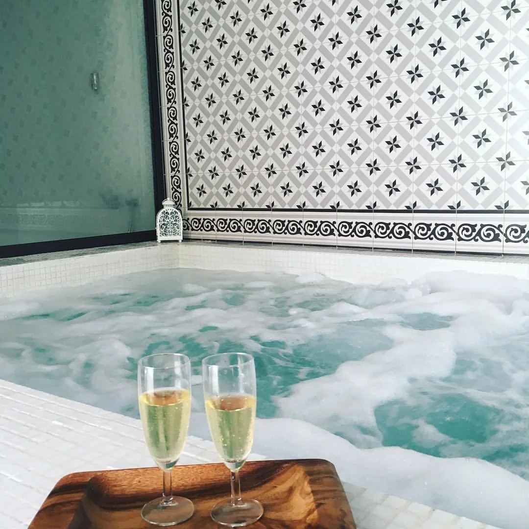 Dos copas de vino espumoso sobre una bandeja de madera junto a una piscina con burbujas, en un espacio decorado con azulejos blancos y patrones en blanco y negro.