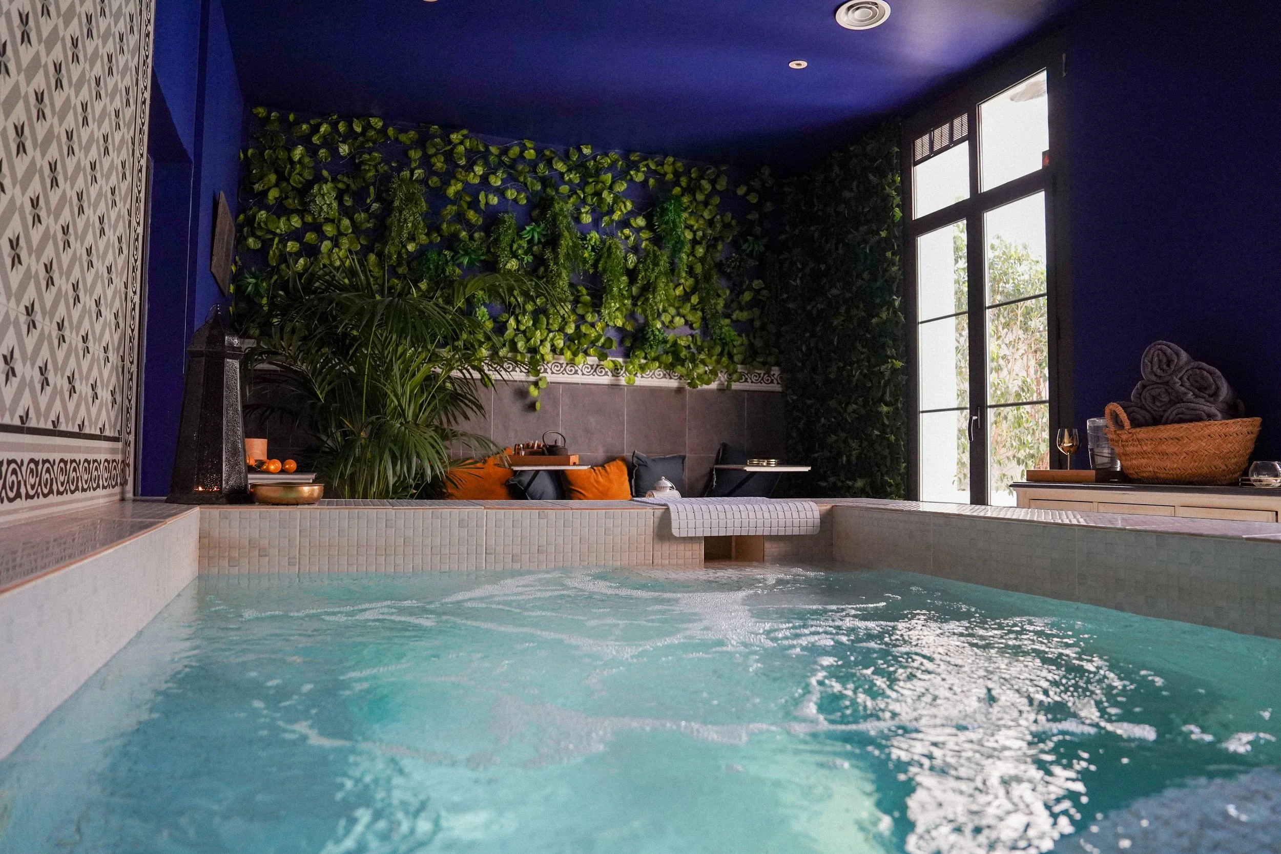 Una piscina cubierta con paredes verdes y ventanas grandes que dejan entrar luz natural, decorada con cojines en el borde y plantas tropicales.