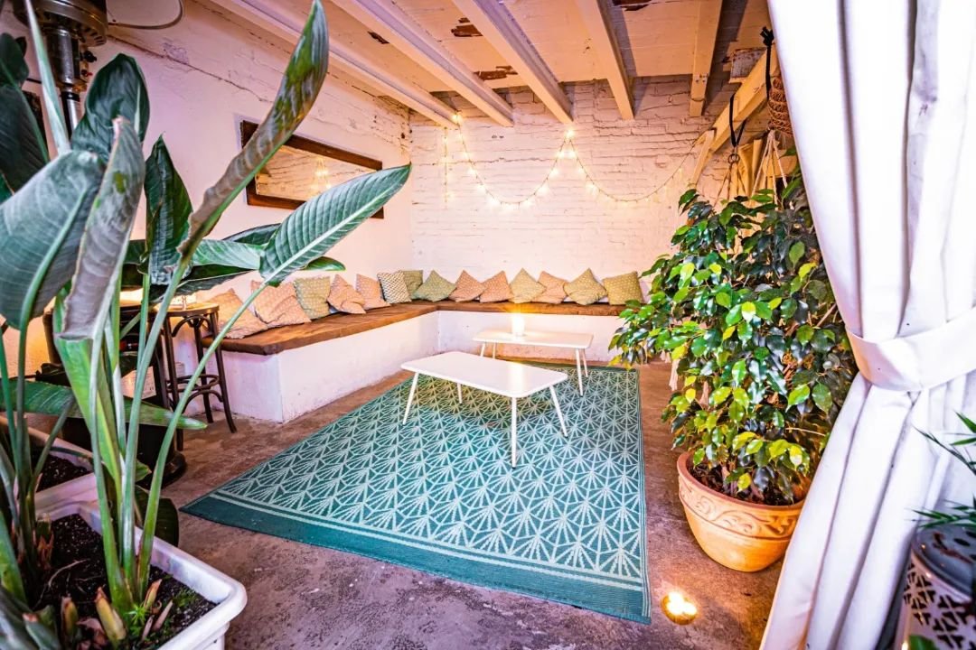 Cozy indoor lounge area with a corner bench, decorative cushions, plants, white tables, and string lights on brick walls.