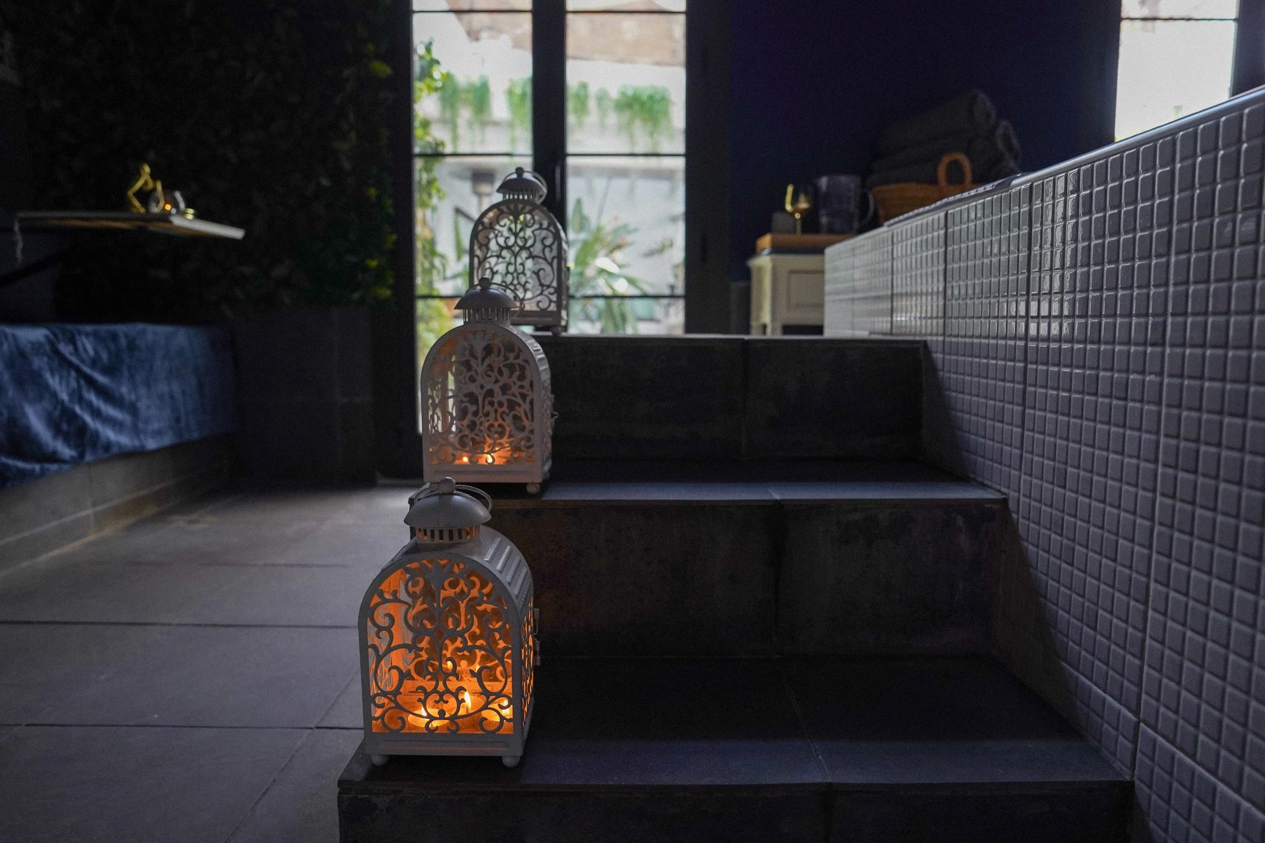 Tres faroles decorativos con luz en una habitación oscura junto a un escalón y una pared de azulejos, con una puerta de cristal al fondo.