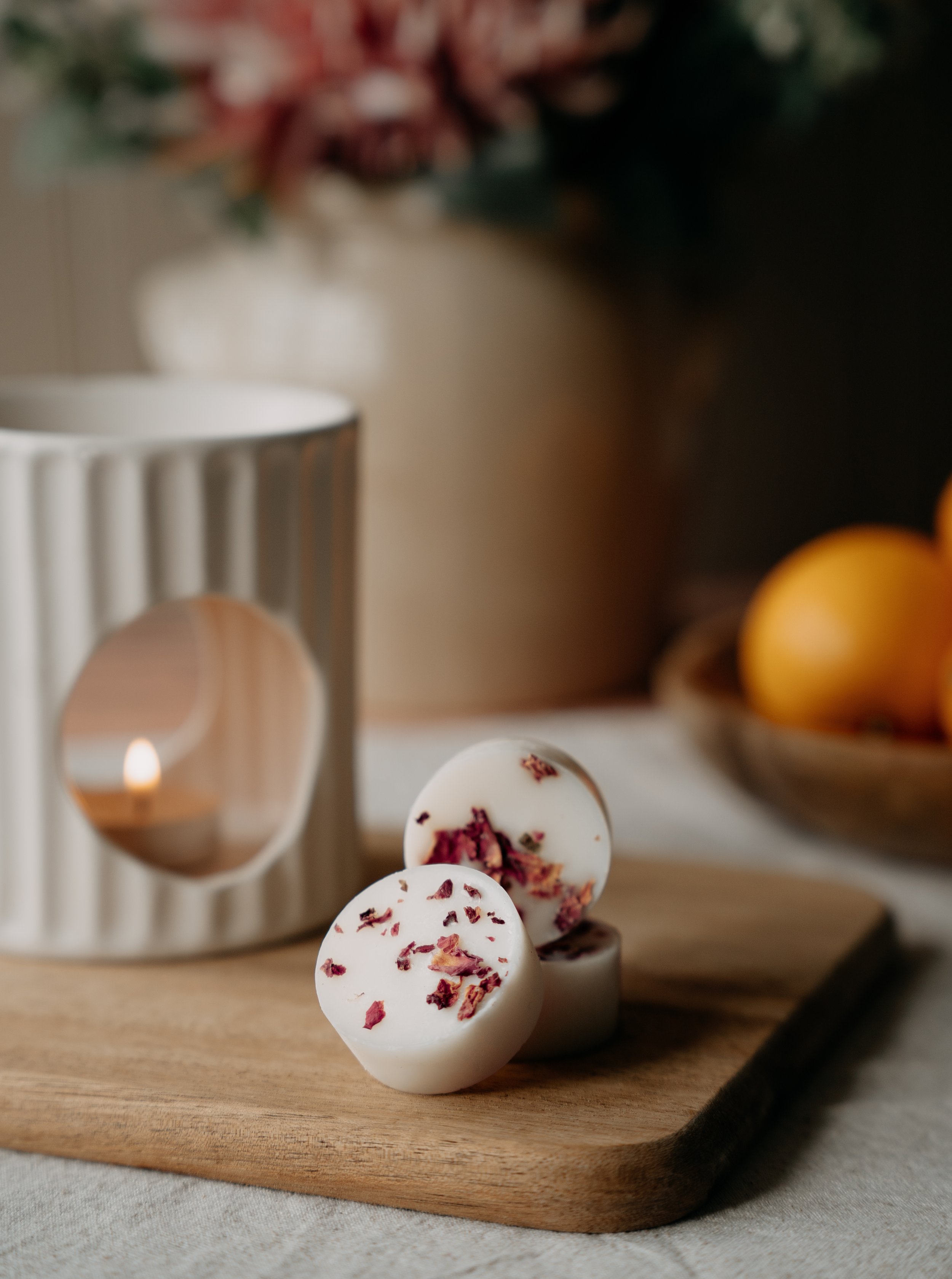 A close-up view of white round bath bombs with pink flower petal decorations on top, placed on a wooden tray with a lit candle and blurred background of a vase with flowers, dark and warm lighting.