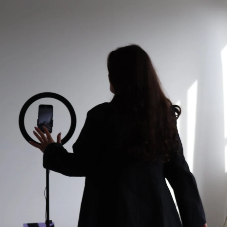 Frau mit langen Haaren macht ein Foto von einem Smartphone, das in einer runden Halterung steckt, in einem dunklen Raum mit Lichtmustern an der Wand.