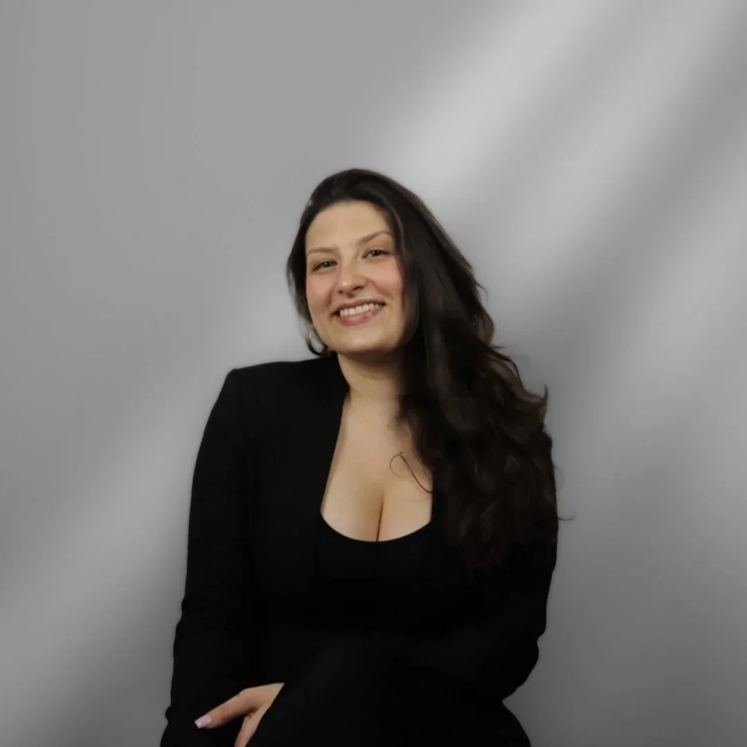 Junge Frau mit dunklen, welligen Haaren trägt schwarze Kleidung und lächelt vor grauem Hintergrund.