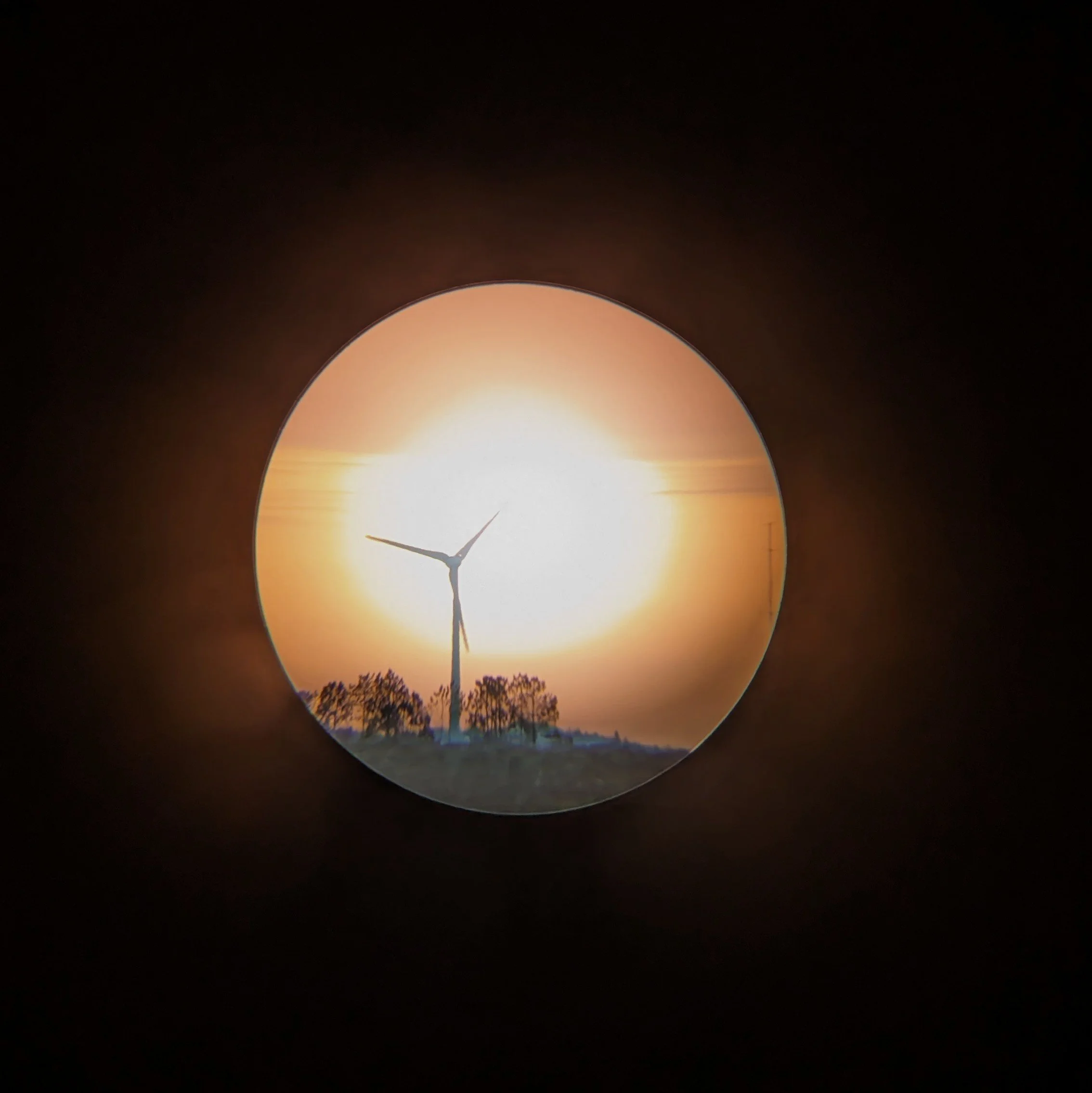 View of a wind turbine silhouetted against a sunset, framed by a circular window or lens.