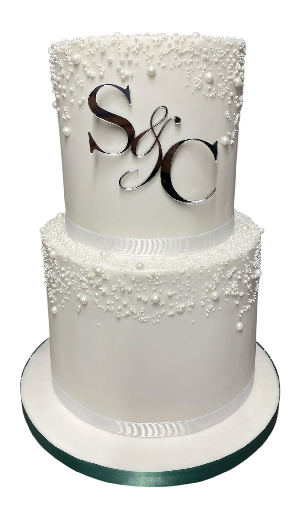 Two-tiered white wedding cake with pearl-like decorations and a silver monogram topper with initials .