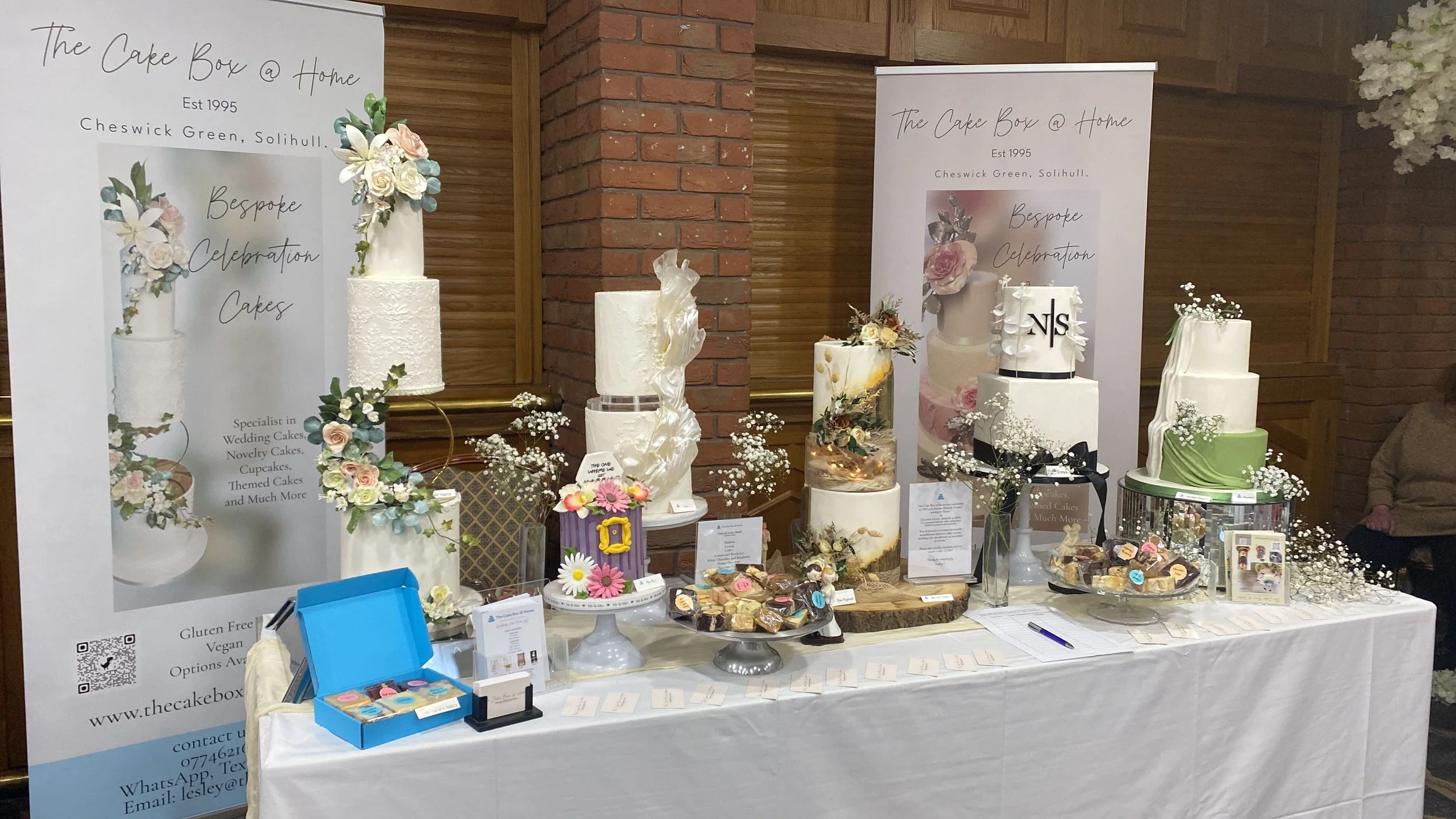 Display table of decorated cakes at a bakery event, with large promotional banners on either side and climbing white tablecloth. Cakes include various styles with floral and abstract designs, surrounded by small flowers, desserts, and promotional materials.