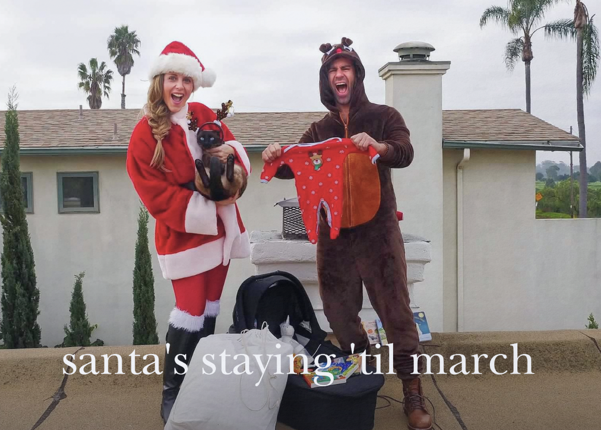Couple in holiday costumes with cat, holding baby clothes, text "santa's staying 'til march"