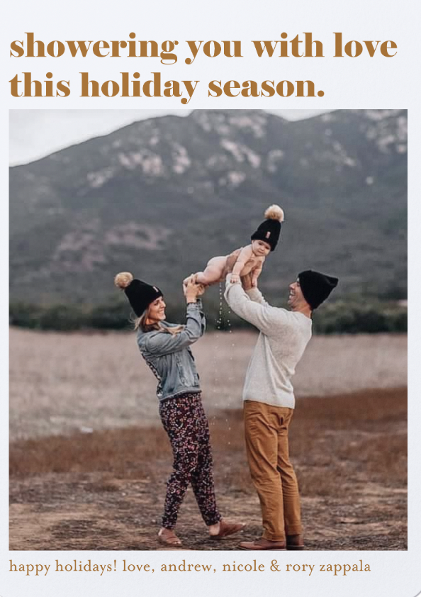 A couple is lifting a child in the air outdoors, with a mountainous backdrop. They are wearing hats with pom-poms. The text reads "showering you with love this holiday season." At the bottom, it says "happy holidays! love, andrew, nicole & rory zappa