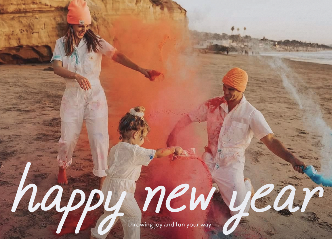Family celebrating New Year on a beach with colored powder, wearing white outfits and orange hats, with text 'happy new year, throwing joy and fun your way.'