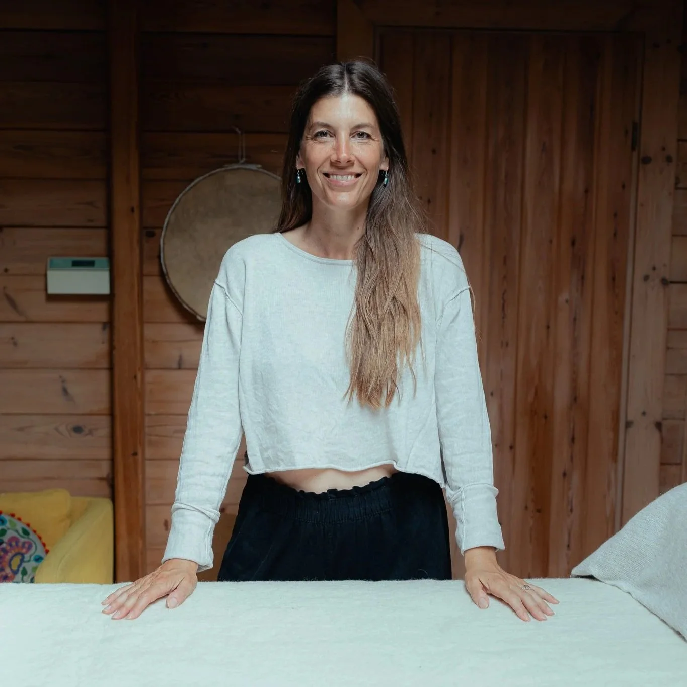A woman with long brown hair, wearing a white cropped long-sleeve shirt and black pants, smiling and resting her hands on a white table in a wooden room.