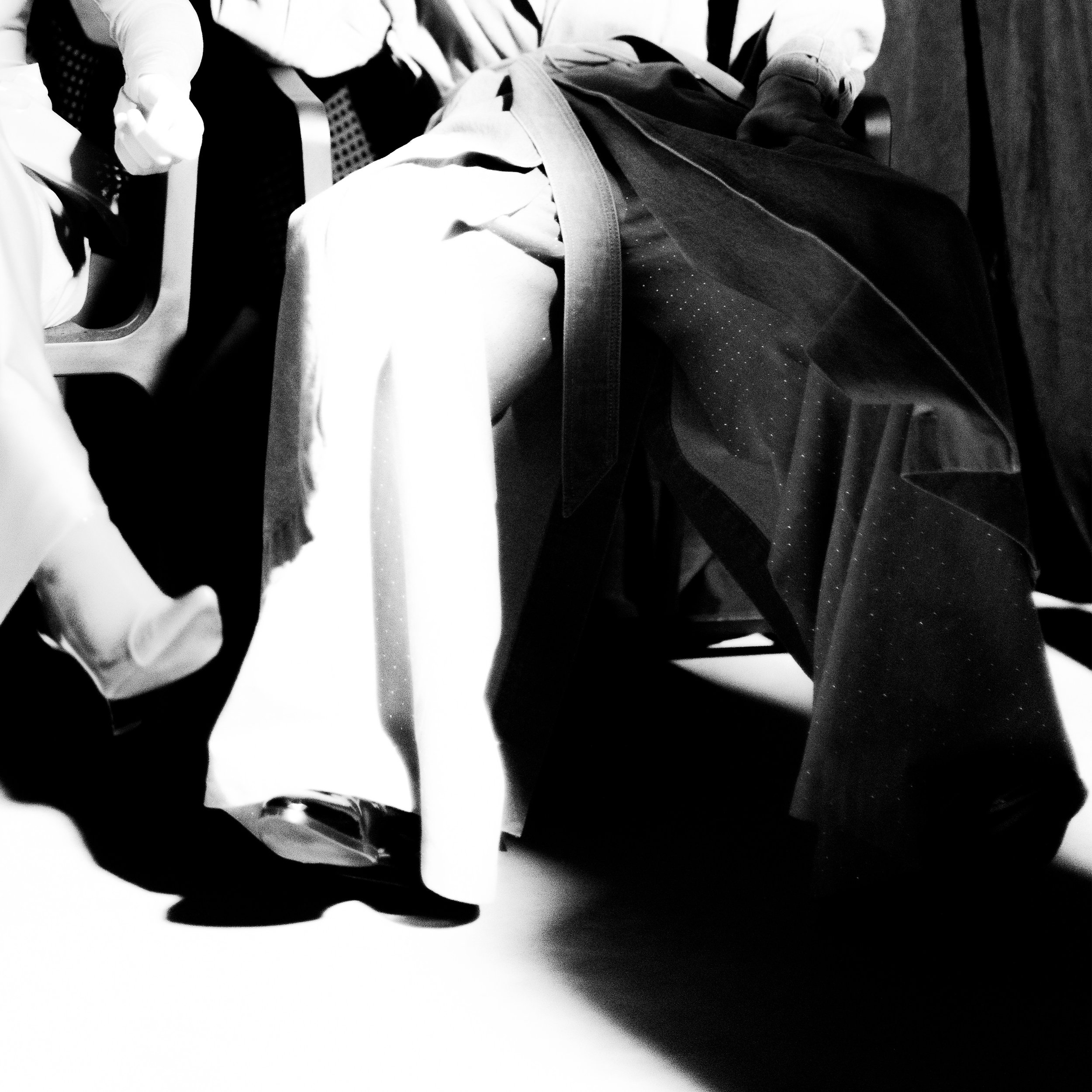 Black and white photo of three seated people, focusing on their lower bodies, wearing different clothing styles including a dress, trousers, and boots.