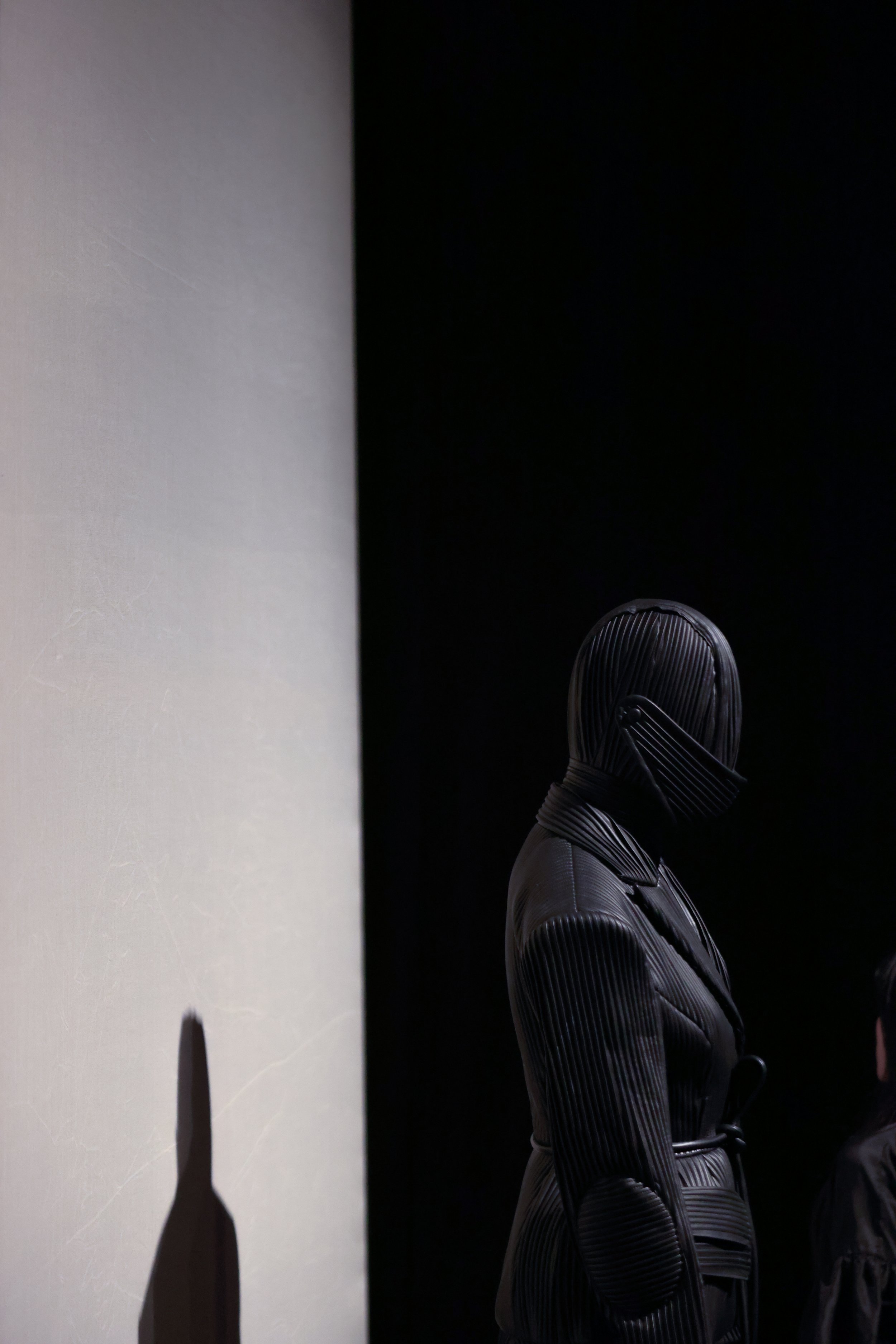 Silhouette of a mannequin or person in a dark, ribbed suit with a full-face covering, standing in front of a gray wall.