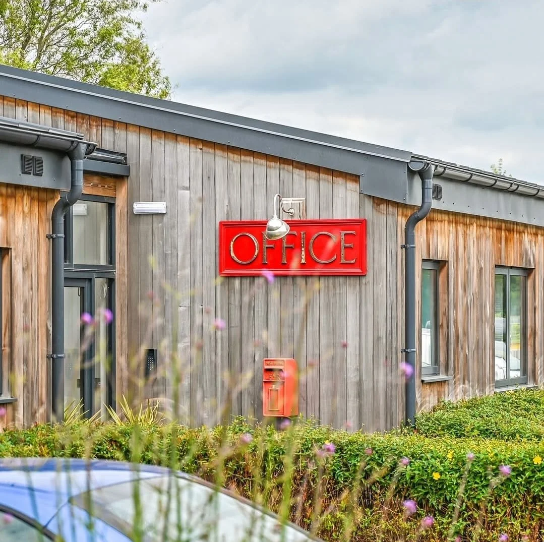 Exterior of a modern building with a red sign that says 'OFFICE' mounted above a window, surrounded by greenery and flowering plants.