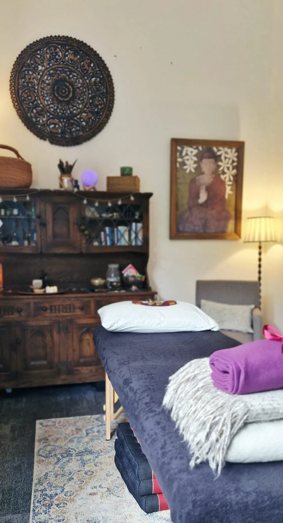 A massage therapy room with a massage table covered in a dark sheet, with a pillow at the head and folded purple towel at the foot, a wooden cabinet with various objects, a framed artwork on the wall, a lamp, and a decorative wall hanging.