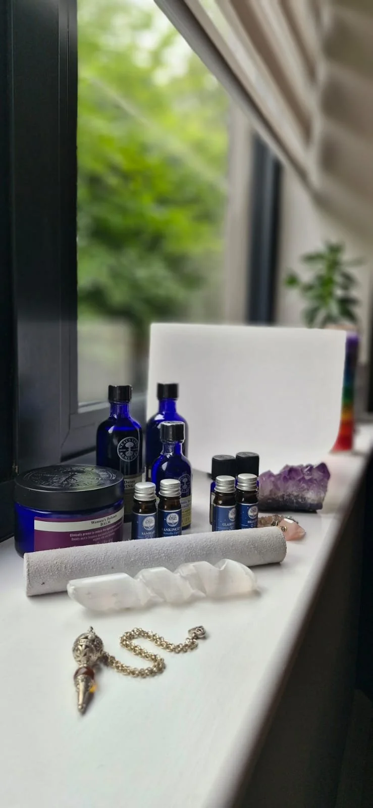 Collection of essential oils, crystals, a white candle, and jewelry on a windowsill with a view of green trees outside.