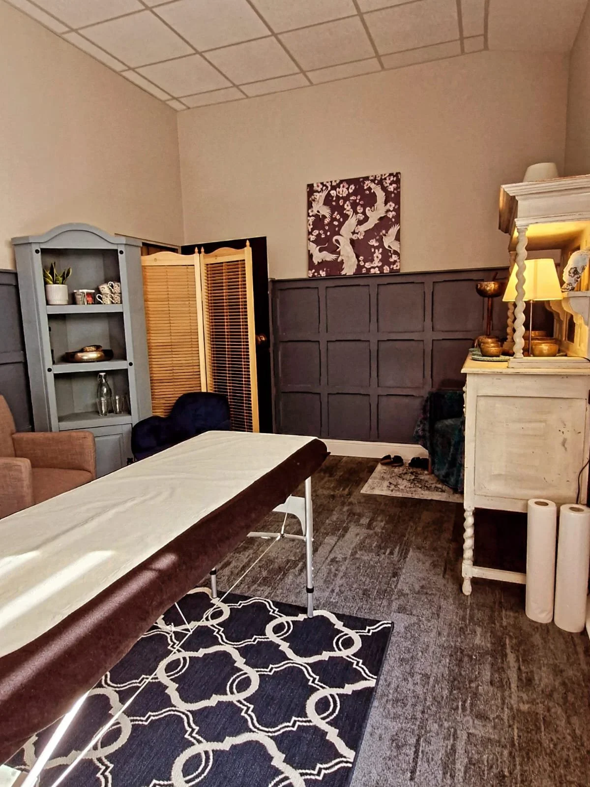 Room with massage table, bookshelf, chair, and decorative artwork on wall.