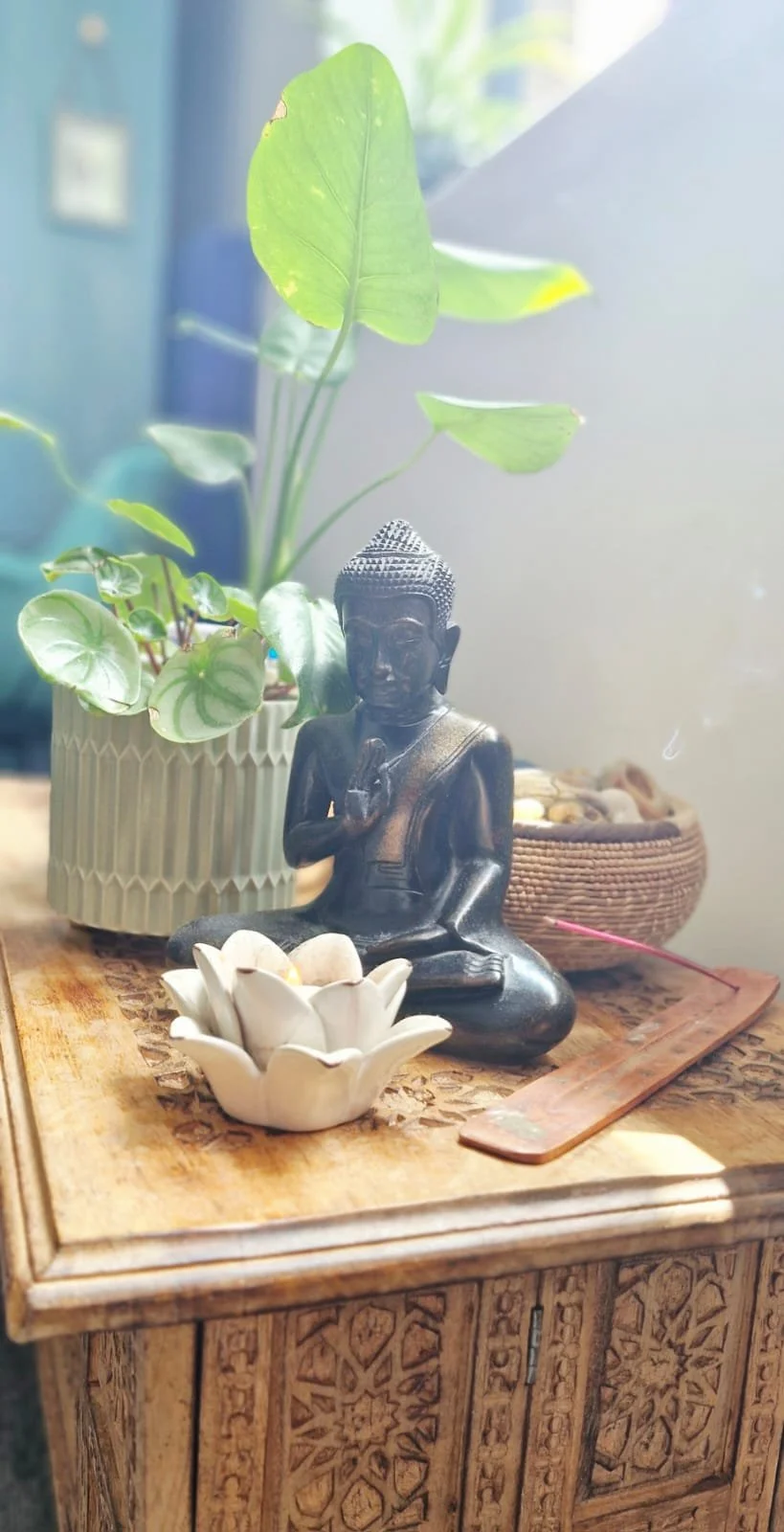 A small black Buddha statue with one hand raised, seated on a carved wooden table, next to a white ceramic flower-shaped bowl with stones, a potted plant with large green leaves, and a basket filled with stones or shells. The background shows a blue