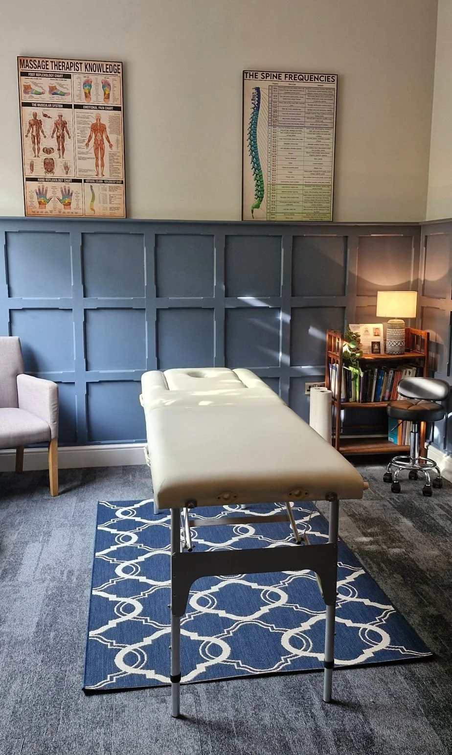Chiropractic or massage therapy treatment room with adjustable massage table, beige upholstered armchair, wooden bookshelf with books and plants, small table lamp, and informational posters about the spine and massage therapy on the wall.