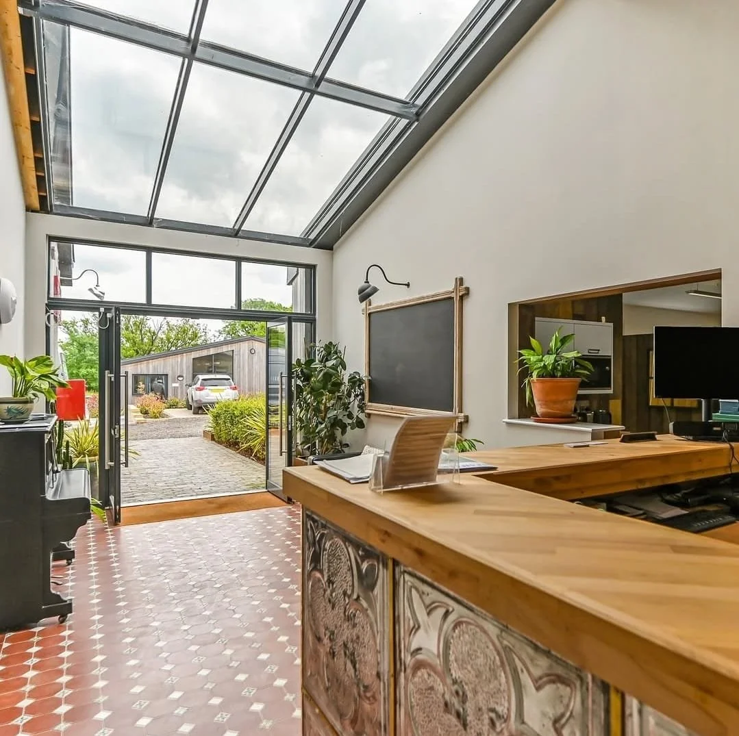 Interior view of a modern reception area with a large glass ceiling, a wooden counter with decorative panels, potted plants, a chalkboard, and an open doorway leading to an outdoor parking lot.