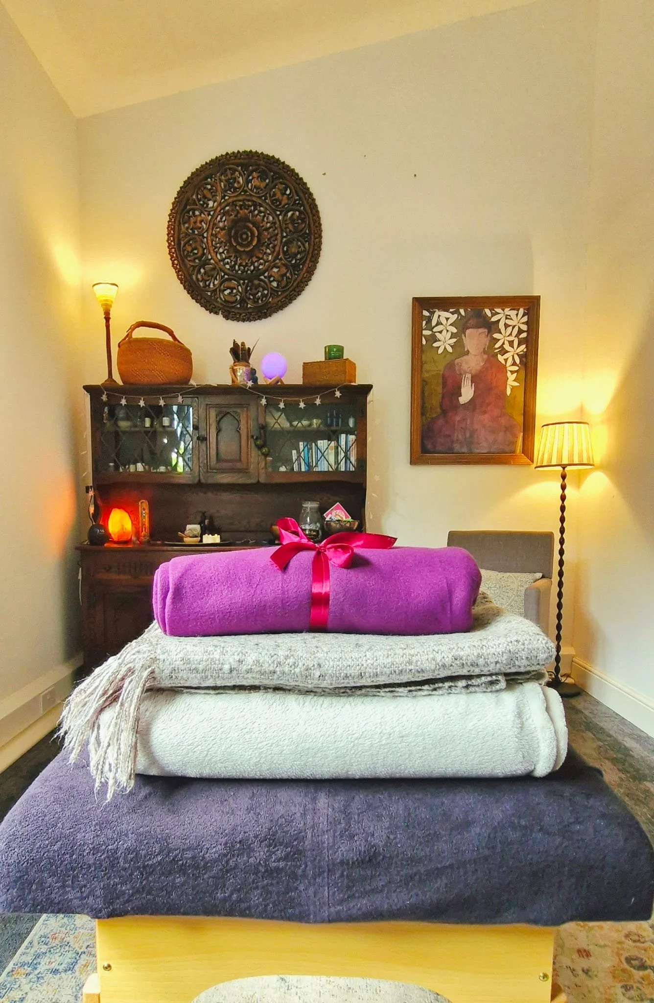 A bedroom with a bed, stacked with blankets and a wrapped purple gift with a red ribbon on top. There is a wooden cabinet with books and decorations, a framed portrait of a woman, and warm lighting from lamps and a wall sconce.
