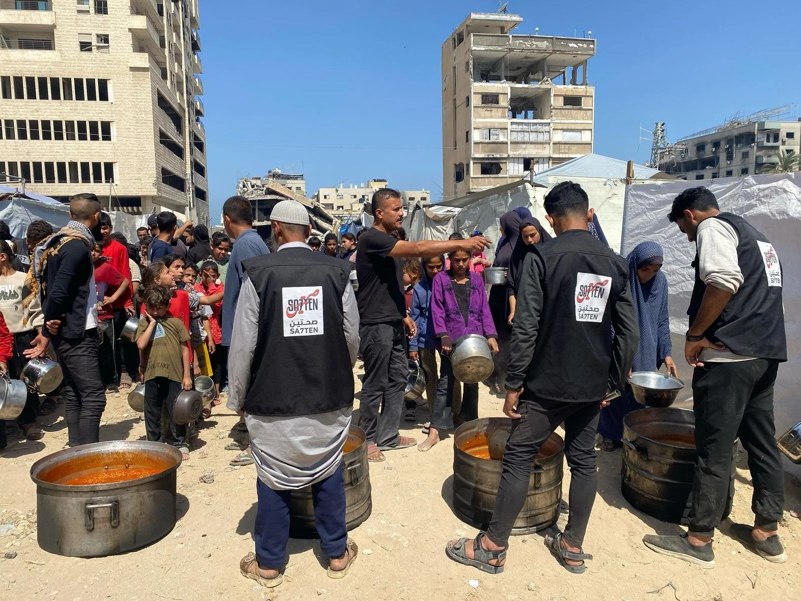 People gather in a crowded outdoor setting with tents and damaged buildings in the background, as some individuals serve hot food from large pots, indicating a humanitarian aid or relief effort.