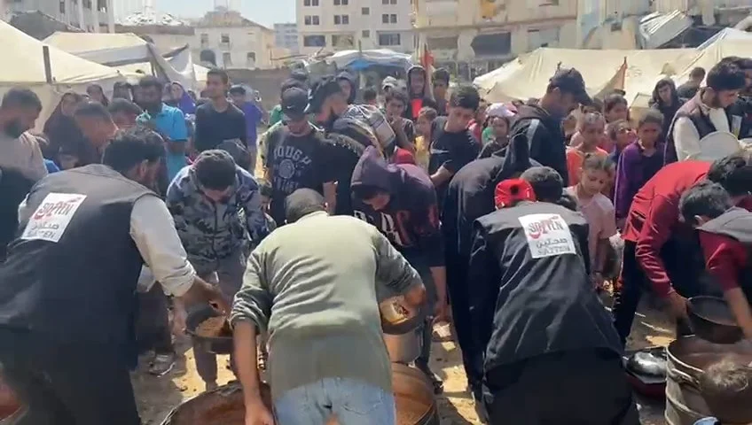 A crowd of people gathered outdoors around volunteers serving food from large containers, amidst tents and urban buildings.