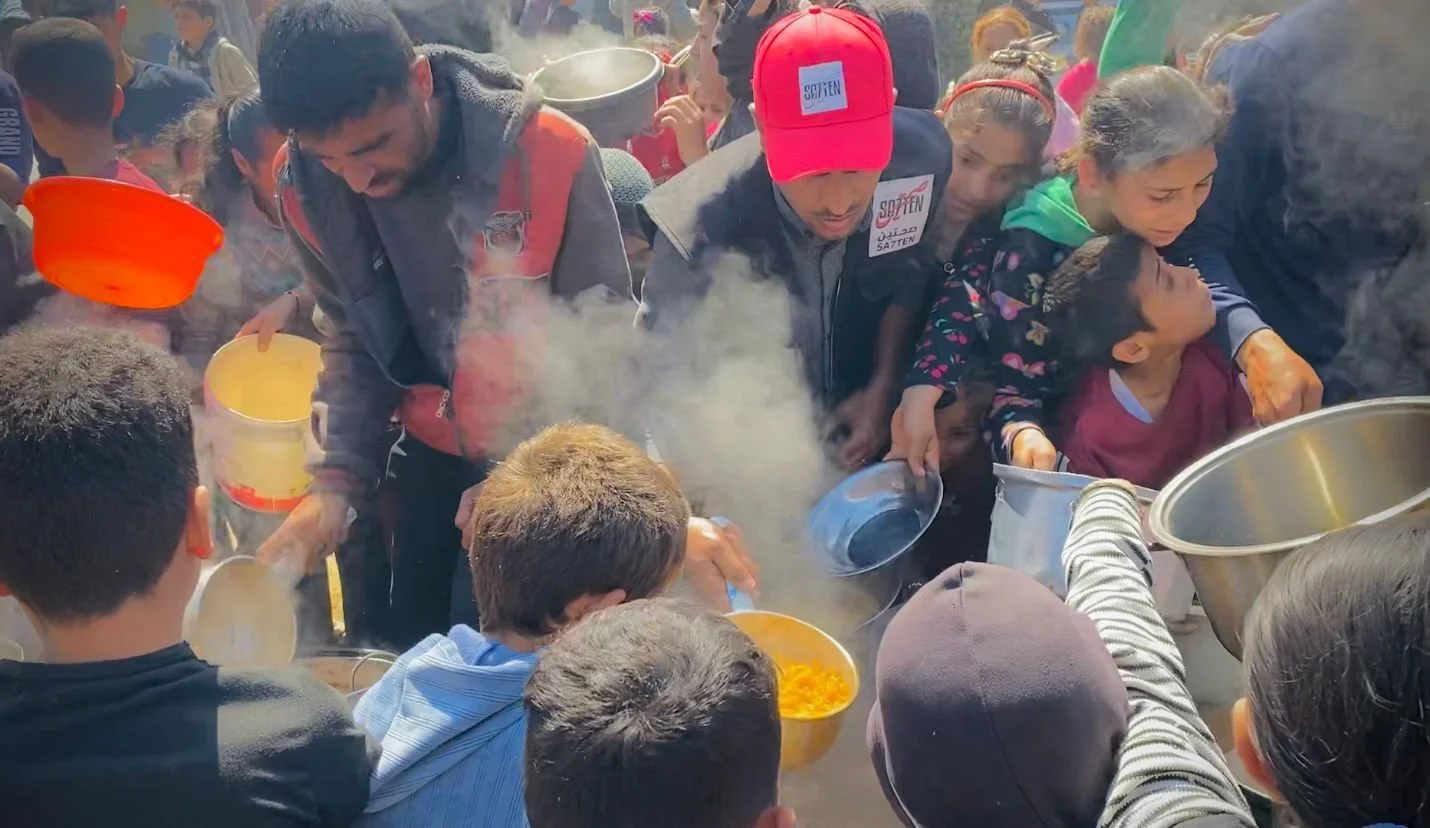 A crowded group of people, including children and adults, gathered around and serving food from large pots, with steam rising, suggesting a community meal or aid distribution.