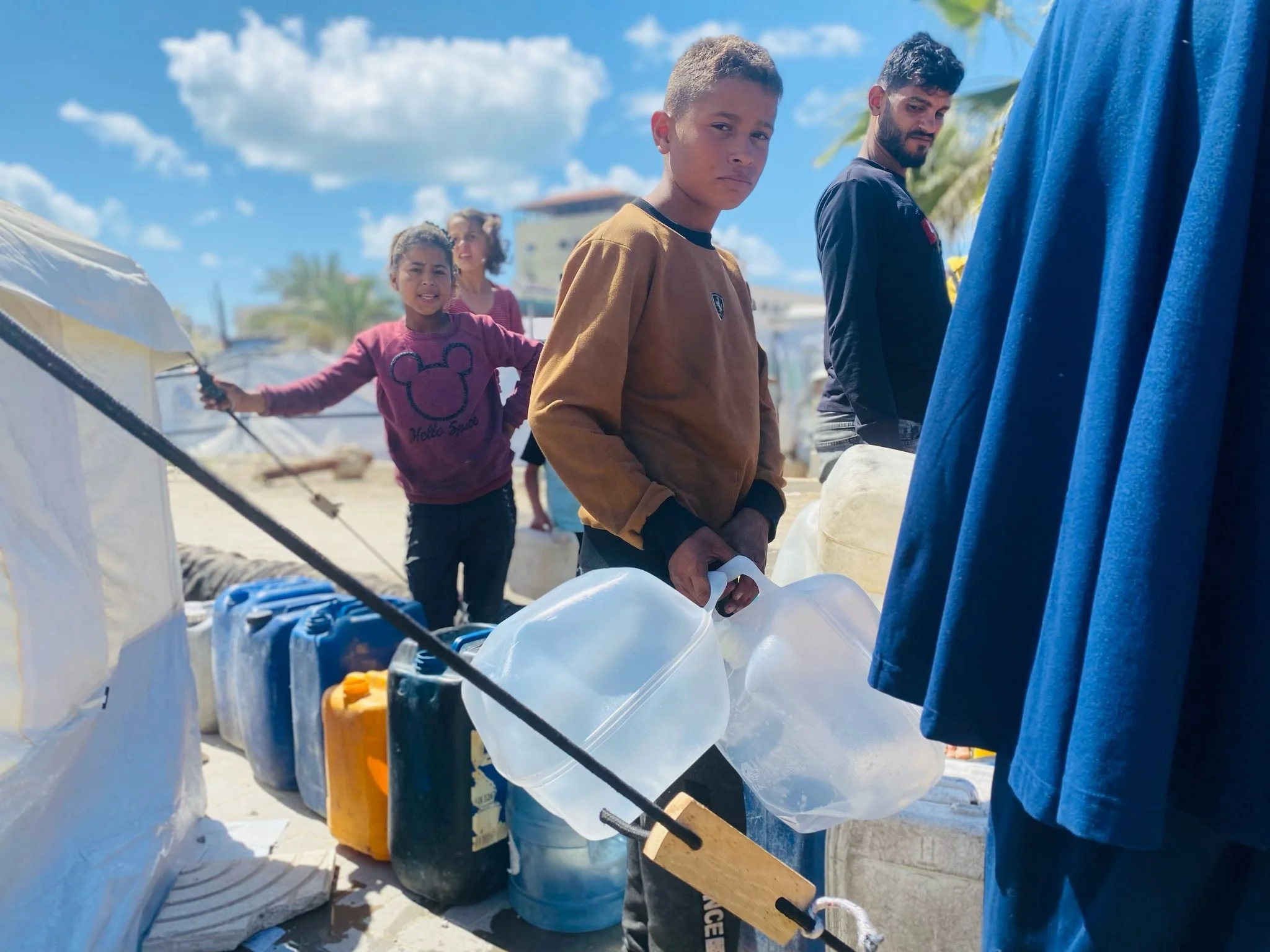 Children and an adult at a refugee camp with supplies, holding plastic containers; blue sky, tents, and palm trees in background.