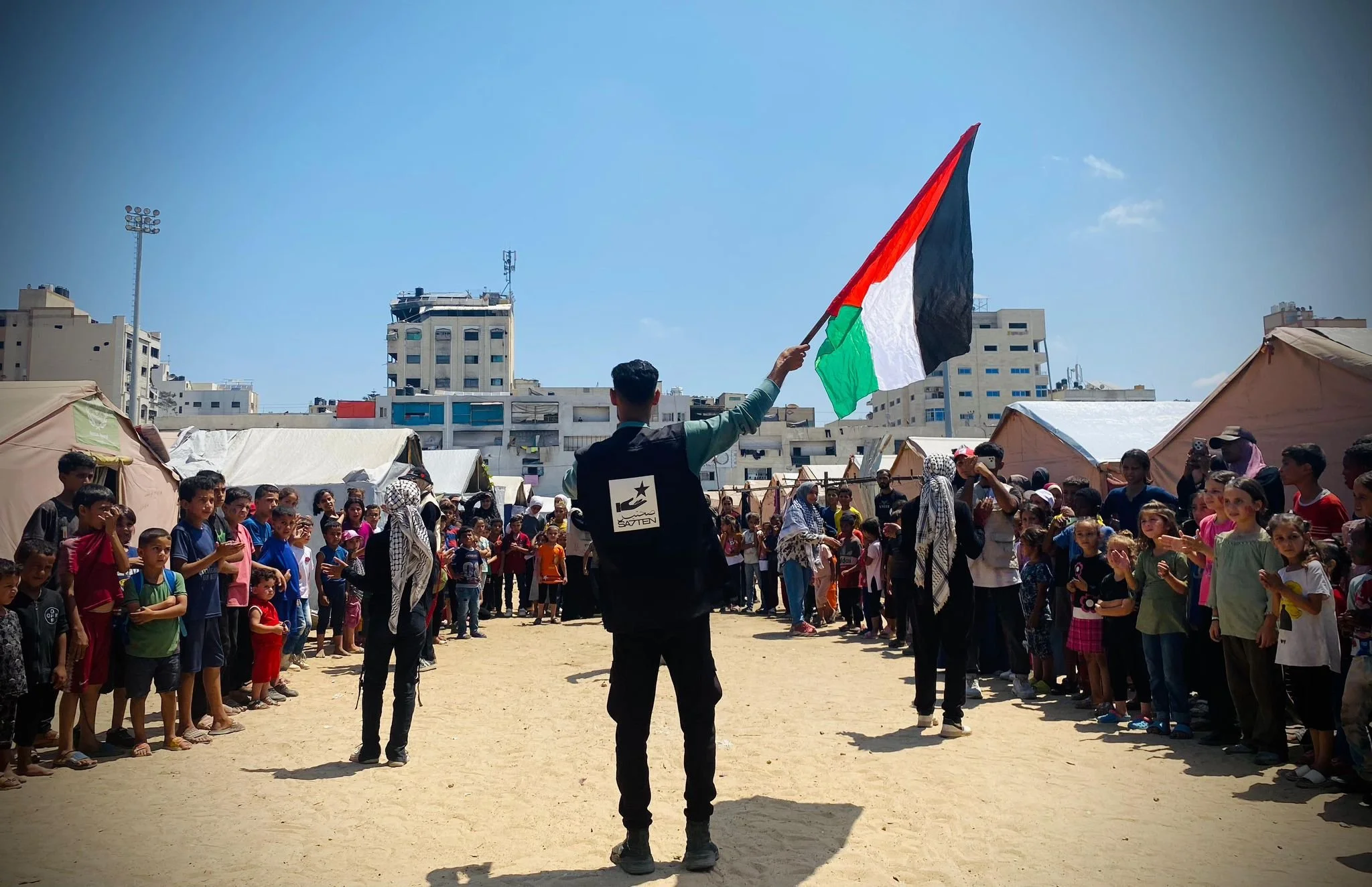 A man holding a Palestinian flag in front of a large group of children and adults gathered in an outdoor area with tents and high-rise buildings in the background.