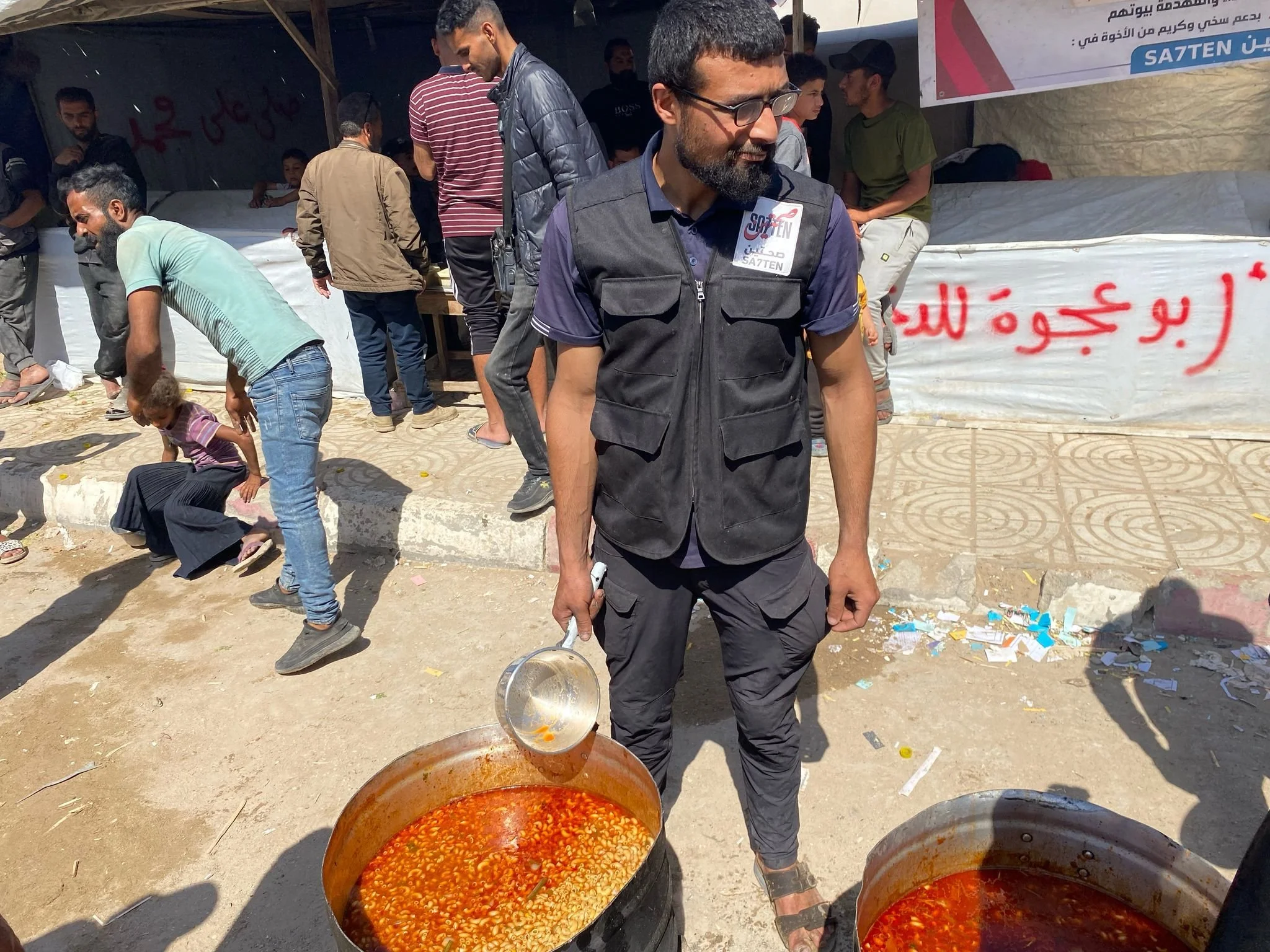 A man wearing glasses and a black vest is standing outdoors, holding a ladle over large pots of red soup. Several people, including children and adults, are gathered behind him, some looking at the camera and others engaged in activities. There is a 