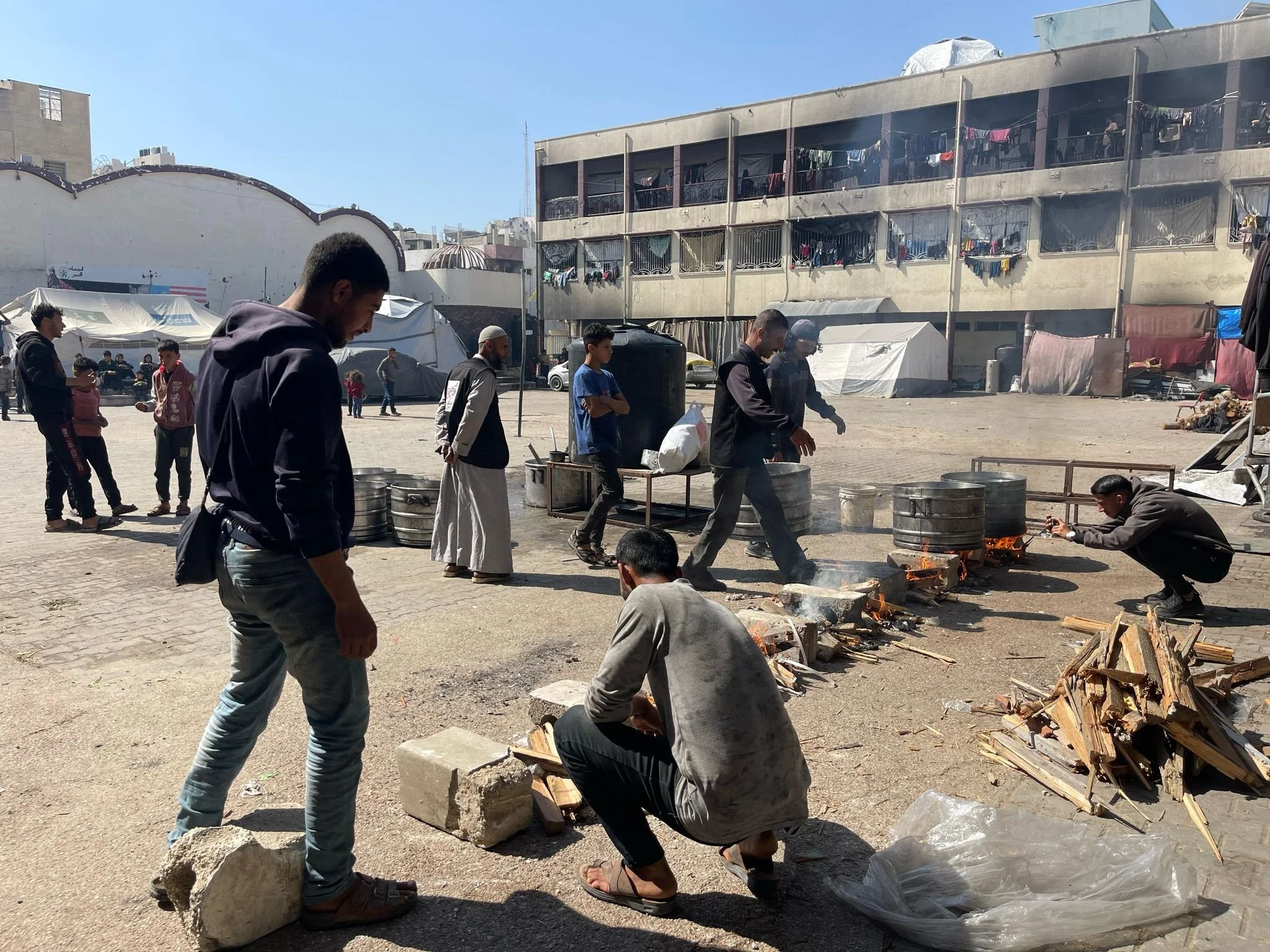 People gathered around outdoor cooking fires with large pots and wood logs in a communal area near tents and a residential building.