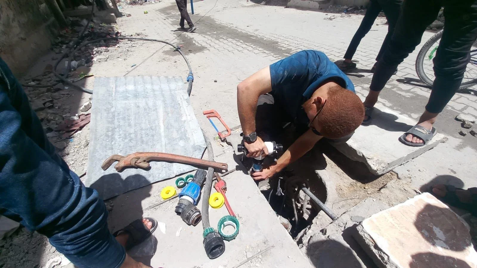 A man working on a water pipe in an outdoor construction site with various tools around him, including a pipe wrench, a hacksaw, and pipe fittings.