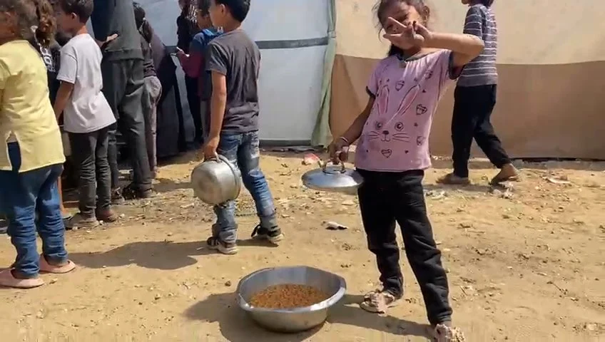 A young girl holds a metal plate and stands next to a large metal bowl filled with food, making a peace sign with her other hand. Several people stand in line behind her, waiting to receive food in an outdoor setting with dirt ground and makeshift te