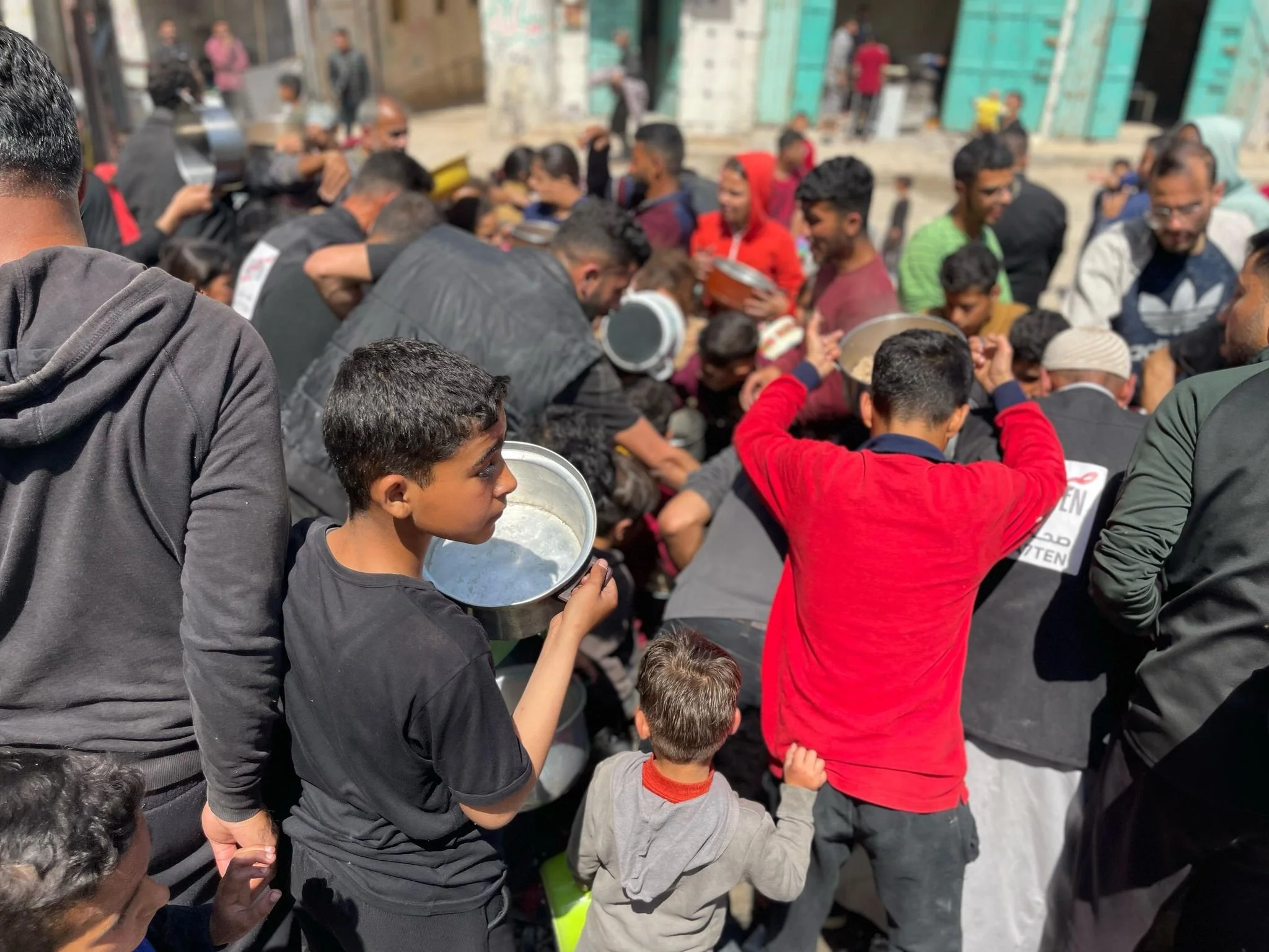 Crowd of people, including children and adults, gathered outdoors, some holding bowls and engaging in a communal activity.