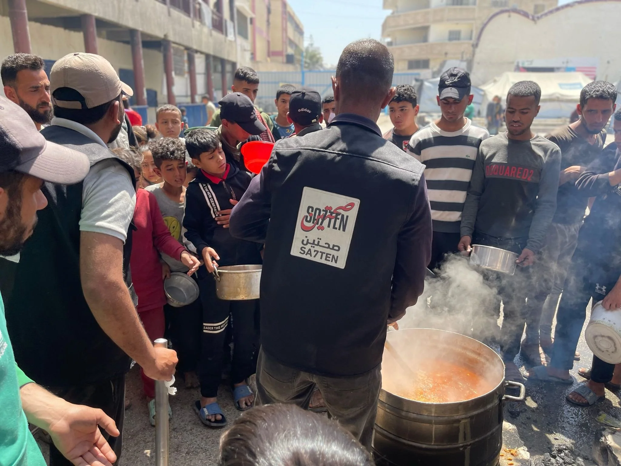 A group of people gathered around a large steaming pot of food on the street, with some holding bowls and looking at the food. The scene appears to be in an outdoor market or street food setting, with buildings and tents in the background.