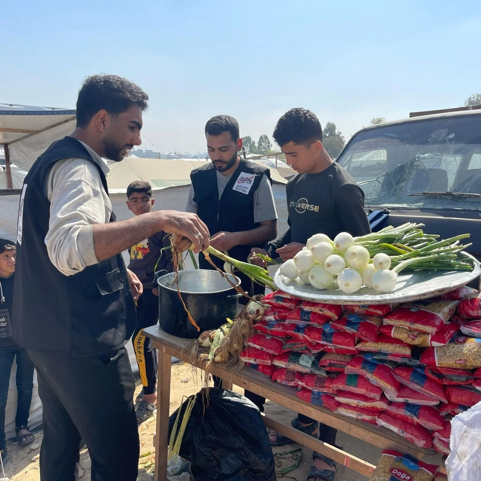 A group of young men at an outdoor market stall with onions, garlic, and packaged food products, some of them preparing or selling produce.