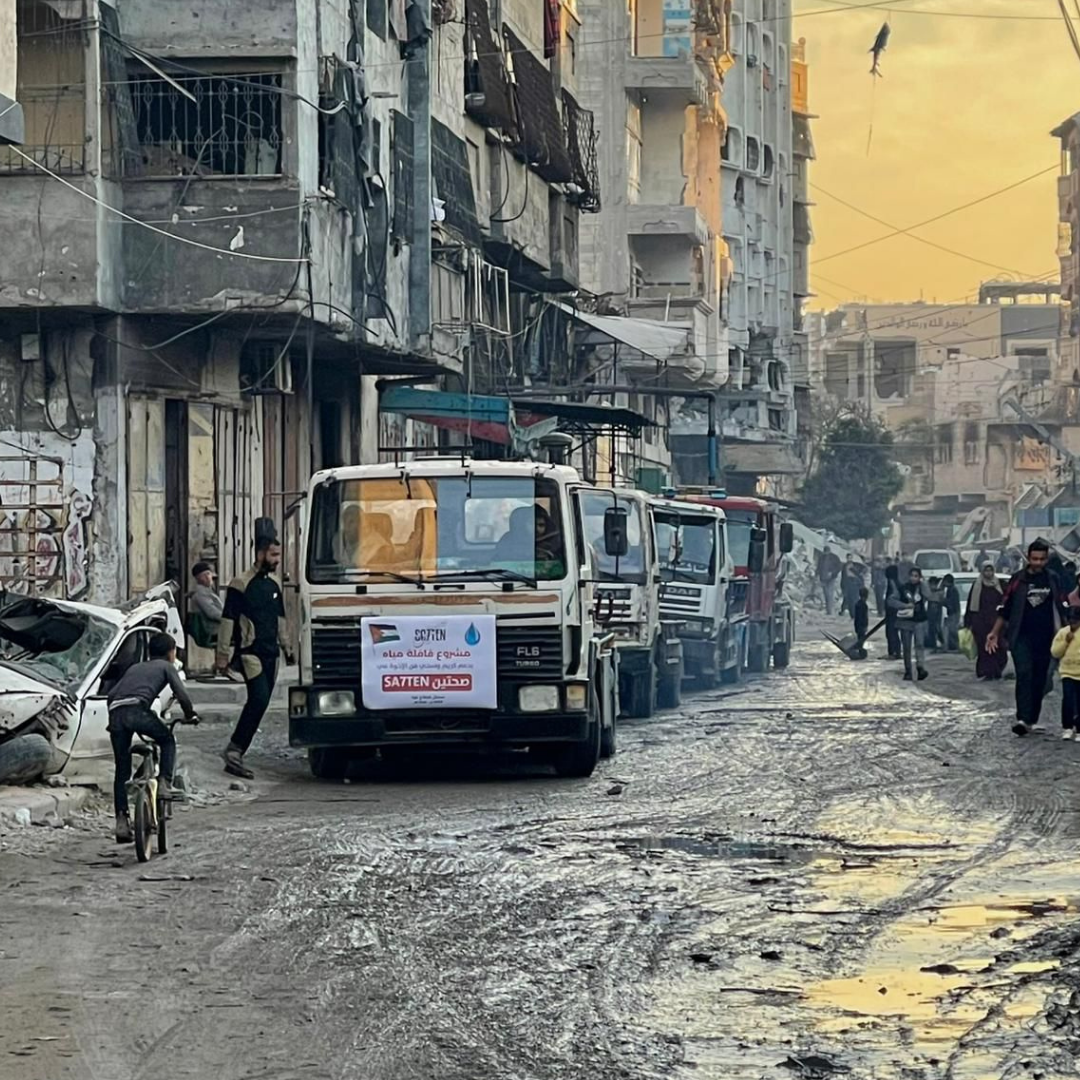 A damaged street with muddy, uneven pavement and damaged buildings. Several trucks and people are visible, with some debris and a partially destroyed vehicle on the left side.