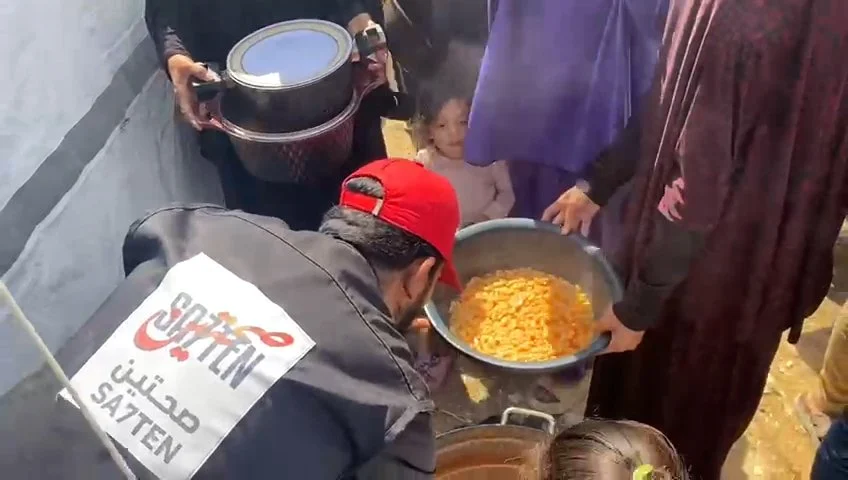 People serving food in a community setting, one person pouring out a large pot of orange-colored food, possibly pasta or rice dish, while others help or observe.