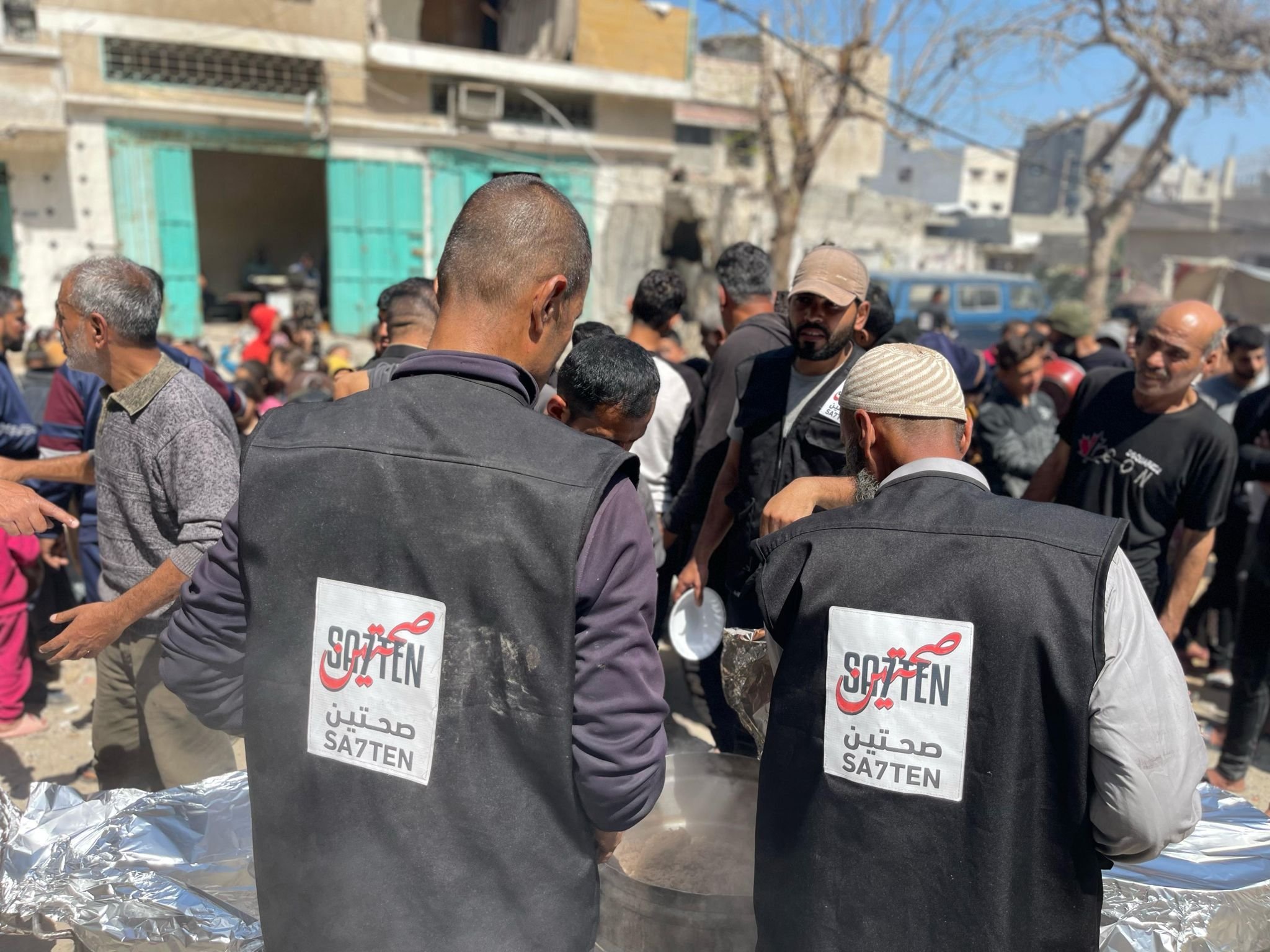 Two men wearing black vests with the word 'SAZTEN' in English and Arabic on the back serve food to a crowd in an outdoor setting with many people and buildings in the background.