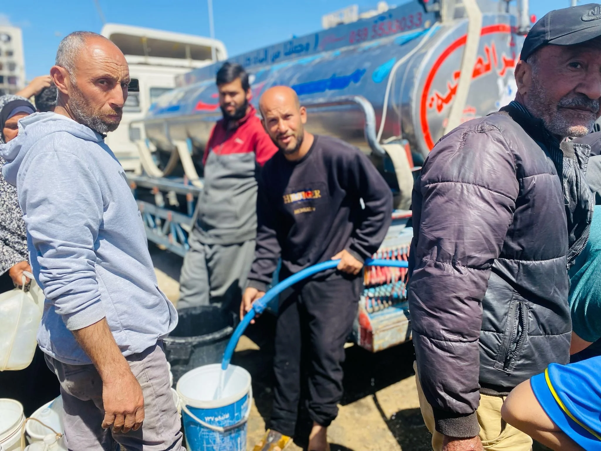Group of men working with water hoses and a tanker truck, outdoors, with bright blue sky.