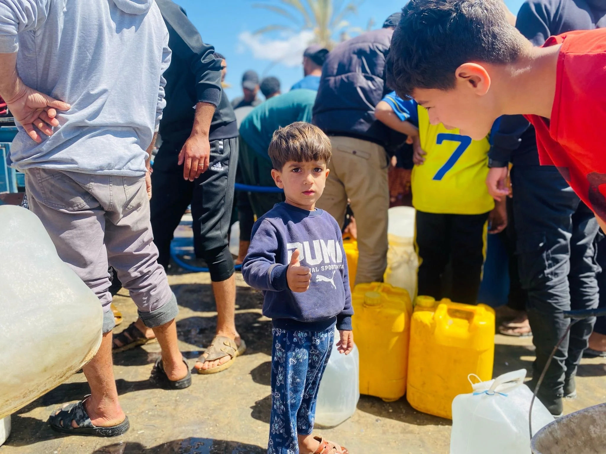 A young boy giving a thumbs up amidst a crowd of people filling yellow and white jerrycans with water.