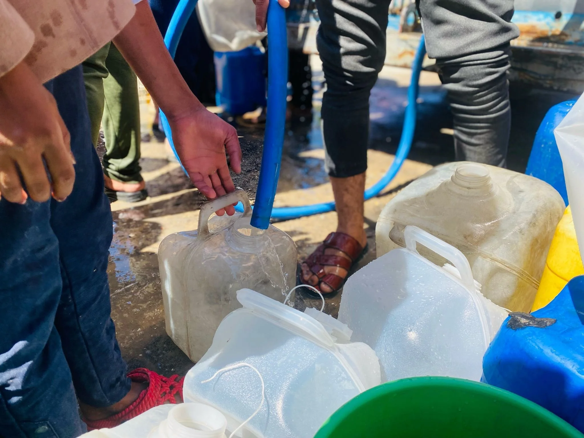 People using plastic jugs to collect water outdoors, with blue hoses connected to the jugs.