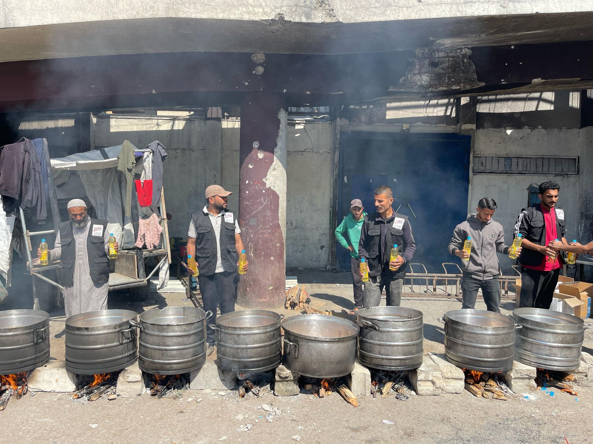 Six men standing outdoors behind large cooking pots over open flames, holding bottles of yellow liquid, in a street setting with a shop or stall in the background.