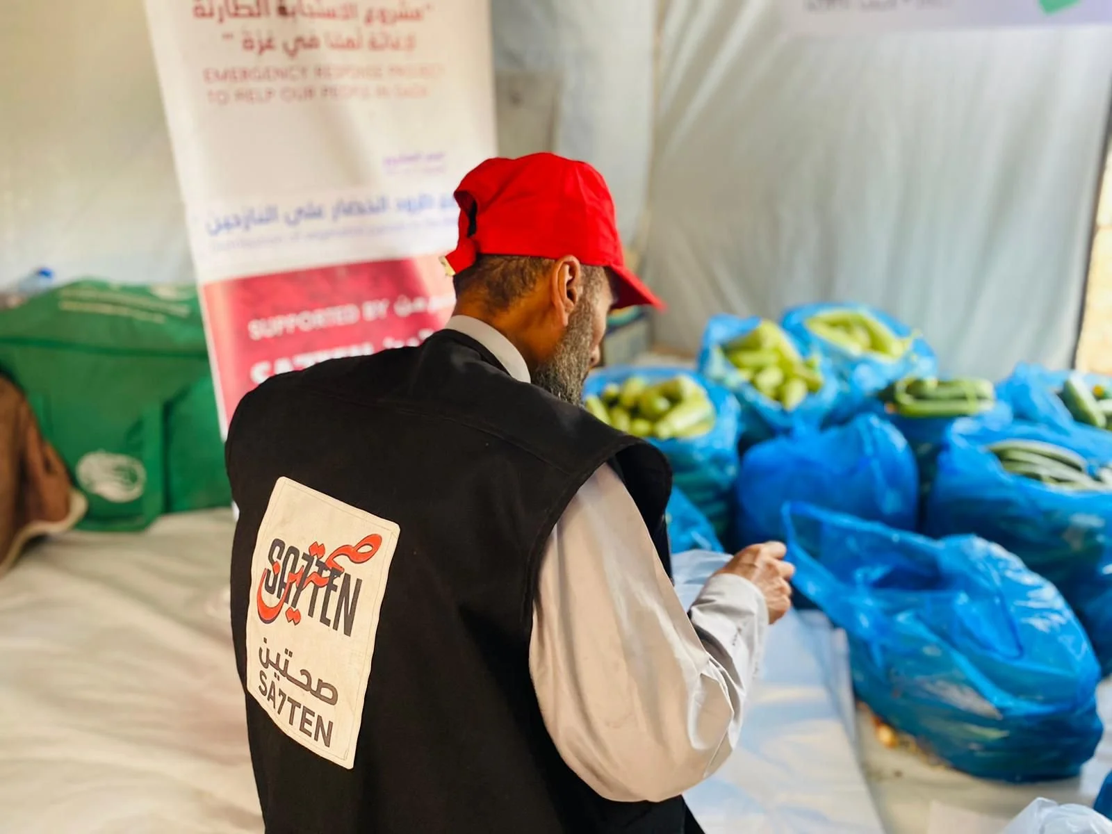 A man wearing a black vest with a SATTEN logo, a white shirt, and a red cap stands near blue plastic bags filled with green vegetables at a humanitarian aid event.