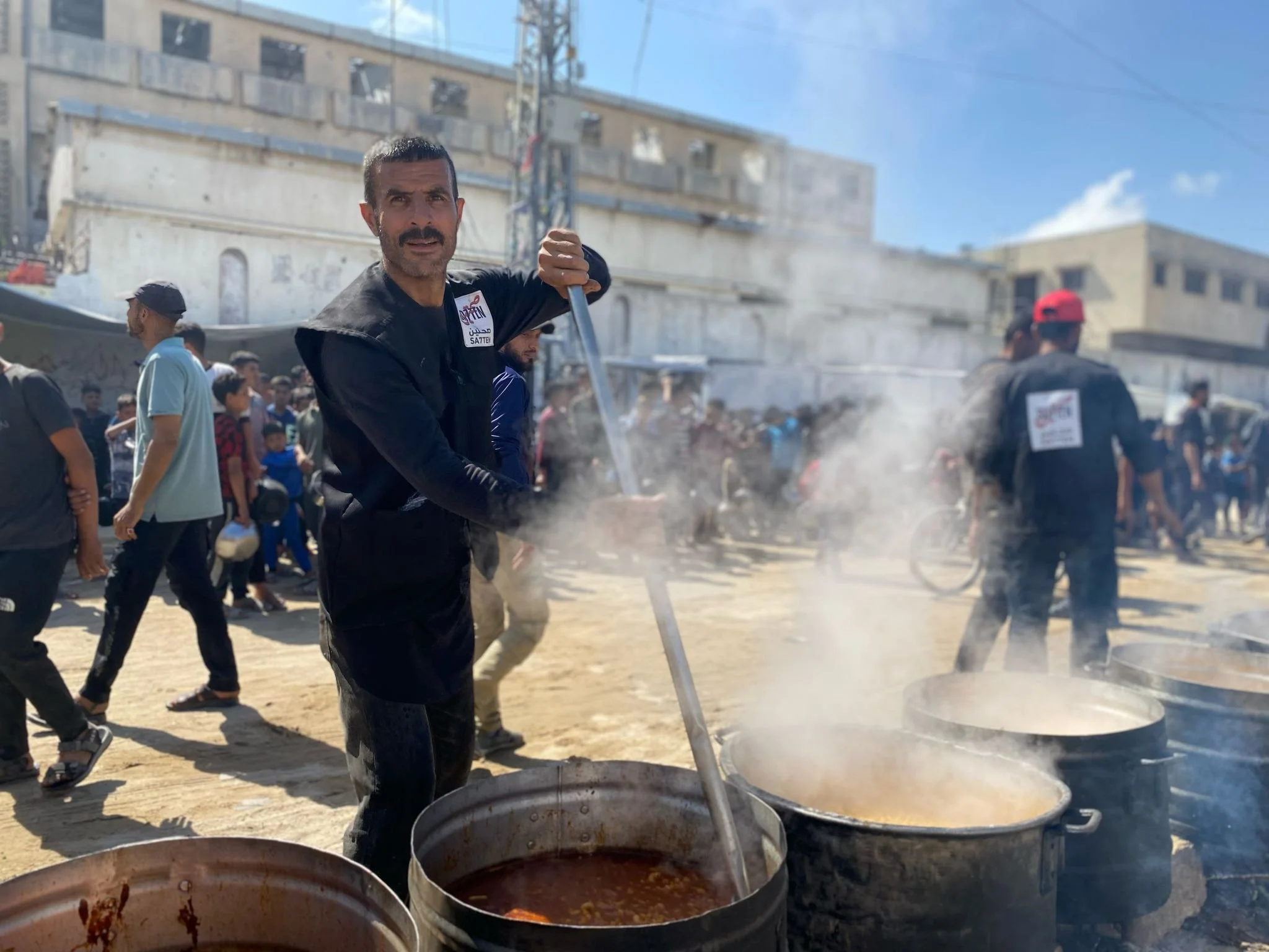 A man stirring large pots of hot food outdoors during a crowded event or gathering.