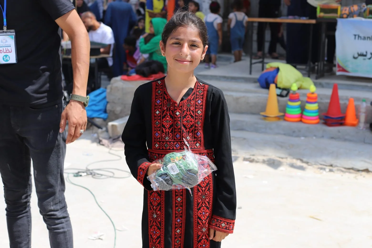 A young girl standing outdoors at an event, holding a plastic bag with a green item inside, wearing a traditional black dress with red embroidery, with a smile on her face.