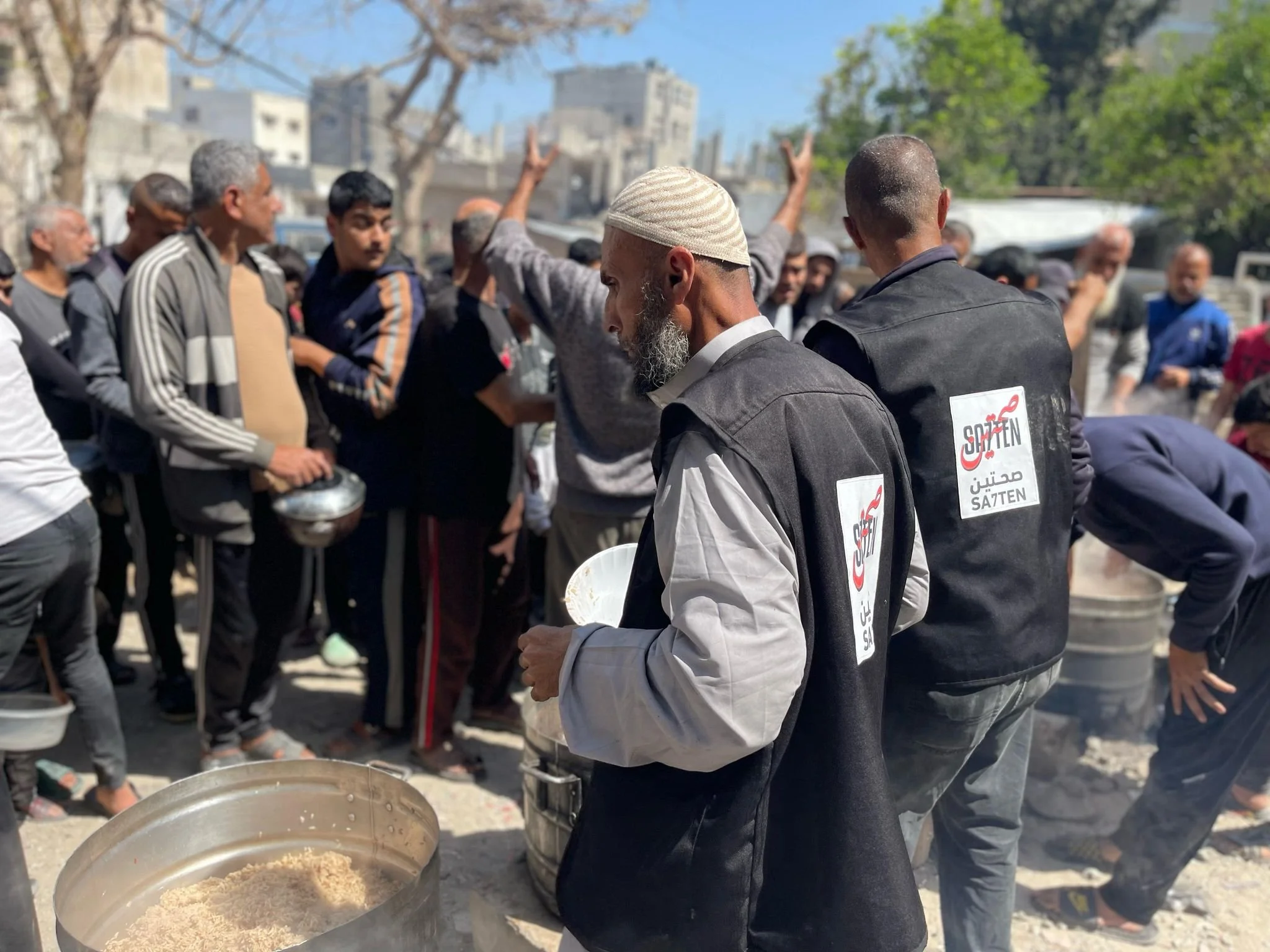 A group of men, some wearing jackets with the logo 'Satlen,' gather outdoors, with some holding bowls of rice, as others stand around in a line for a meal or distribution.
