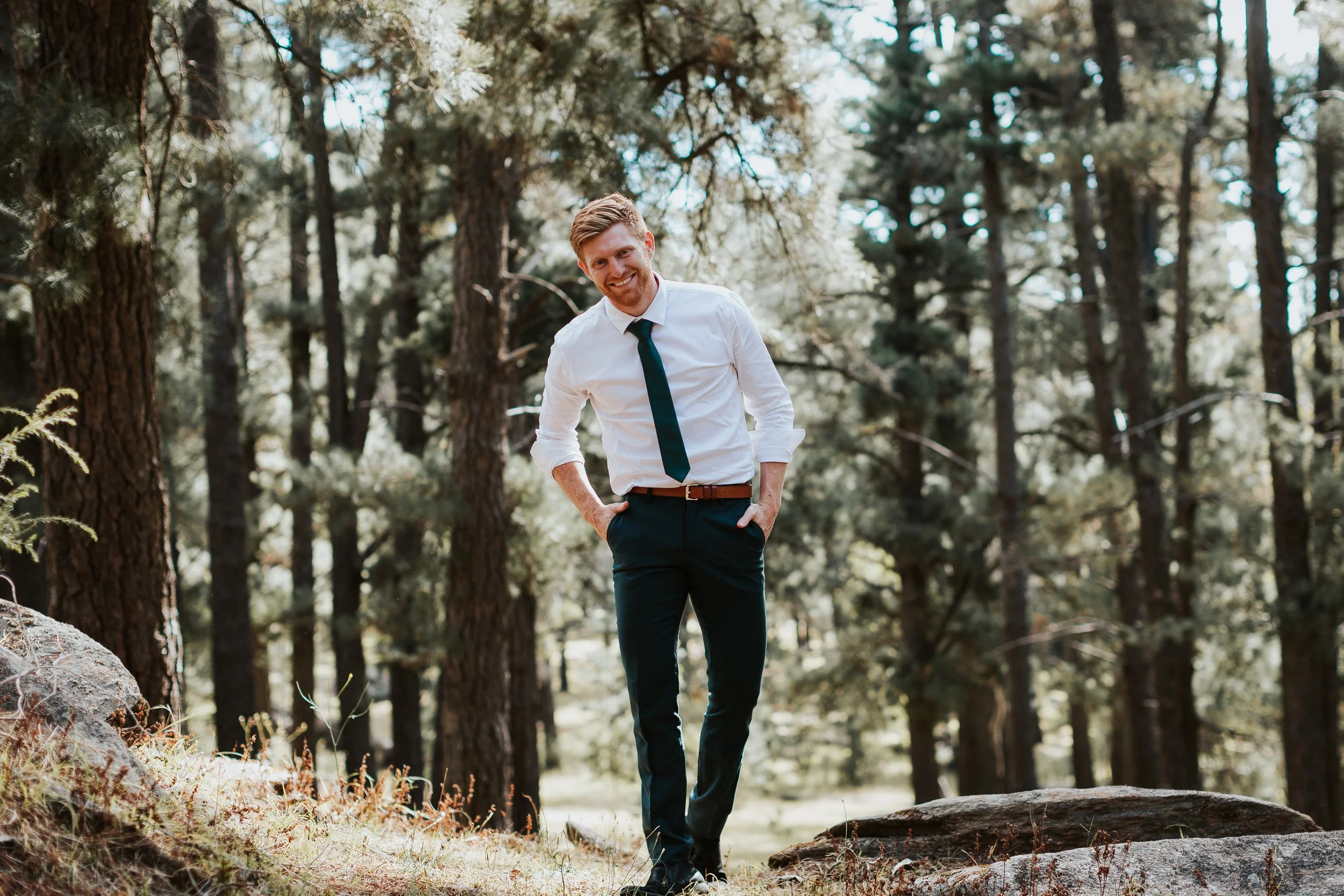 A man in business attire, white shirt and dark tie, walks through a forest with tall trees, smiling and with hands in pockets.